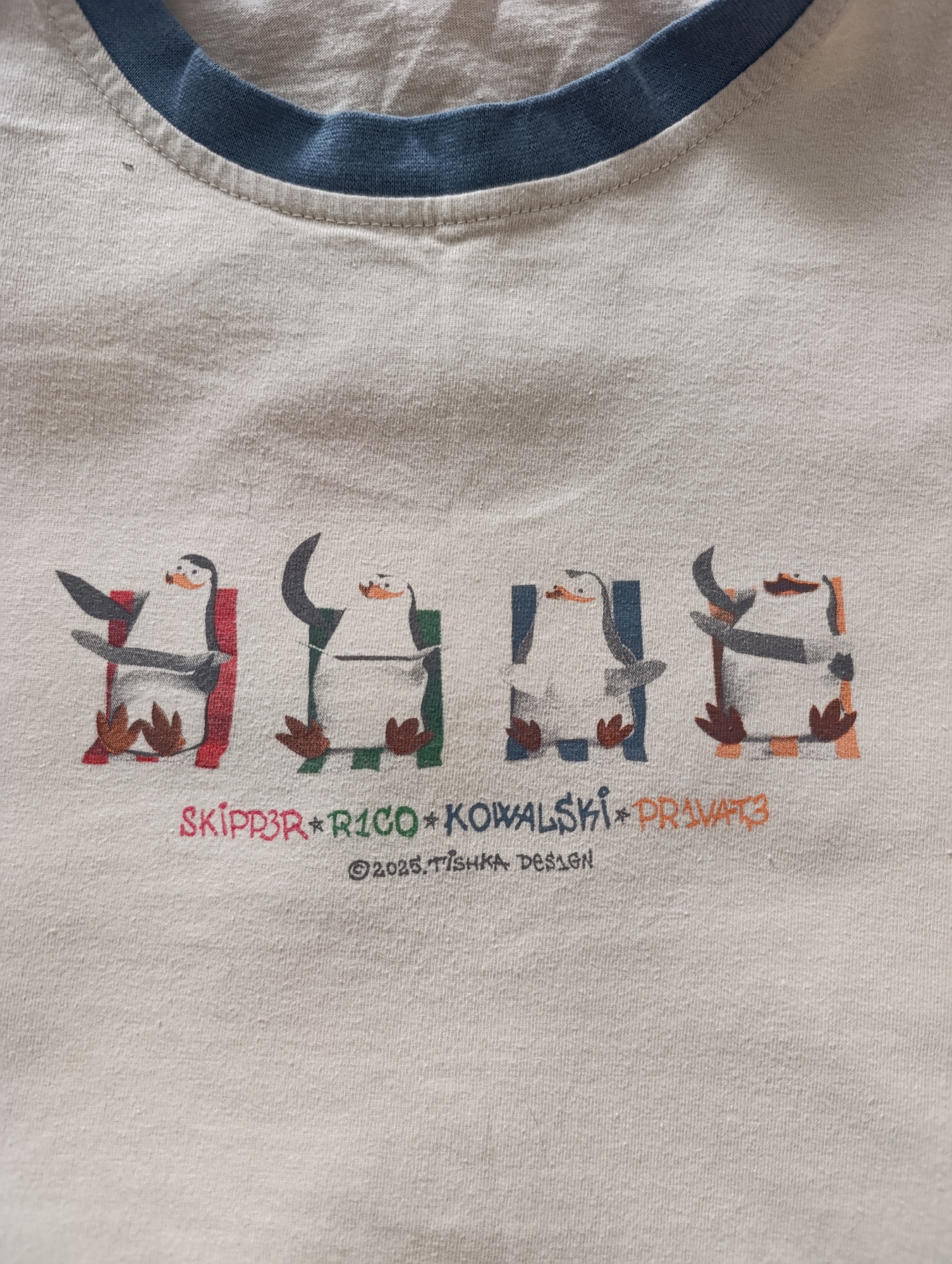 A close-up shot of a white t-shirt with a blue-grey collar. The shirt features a graphic of four penguins stylized in blocky designs with different colored squares behind them. The penguins are labeled "SKIPPER," "RICO," "KOWALSKI," and "PRIVATE," with the names separated by asterisks. Below the penguins, it reads "©2025. TISHKA DESIGN." The text is in a light-colored font. There is no information available about the location being in the city of Chaikovsky, Russia.