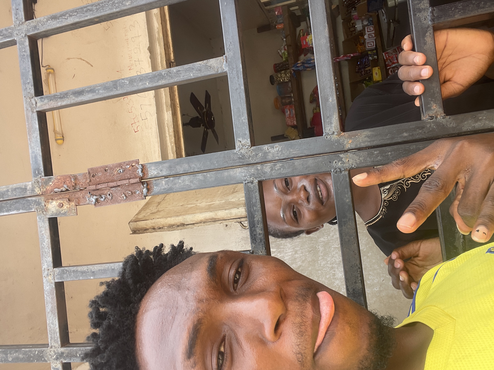 A selfie-style photograph features two dark-skinned individuals looking through a metal barred window. The person in the foreground has short, curly hair and is wearing a yellow shirt. Their face is close to the camera, showing a slight smile. The second person is behind the bars, looking through the window frame toward the camera, wearing a black garment. The metal bars of the window are visible throughout the shot. Behind the individuals, a glimpse of the interior reveals a room with shelves stocked with various items, and a ceiling fan is also visible. A pale wall with writing on it can be seen on the left side of the window. This photograph appears to be taken in Lokoja, Nigeria, given the location information provided.