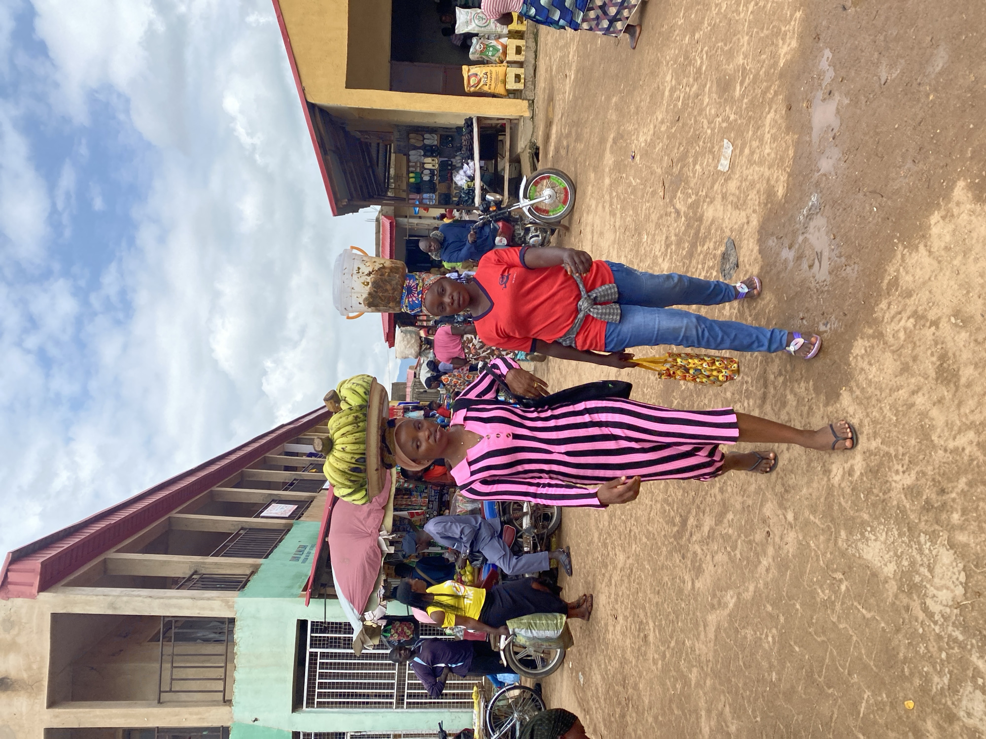 The image depicts a busy outdoor market scene in Kafanchan B, Nigeria. Two women are walking towards the viewer, each carrying a load balanced on their heads. The woman on the left wears a pink and black striped dress and carries a large bunch of bananas on her head. She also carries a black bag over her left shoulder and a shopping bag with yellow smiley faces in her right hand. The woman on the right wears a red t-shirt, blue jeans, and a patterned headscarf. She carries a clear plastic bucket filled with what appears to be food items on her head. A grey and white checkered shirt is tied around her waist.
In the background, other people are visible, some walking and others on motorcycles. Several market stalls are set up, displaying goods such as shoes and bags of grain. Buildings with corrugated metal roofs and open-fronted structures line the market area. The ground is unpaved and appears dusty, with some patches of mud or standing water. The sky is partly cloudy. The overall impression is one of a vibrant, working marketplace.