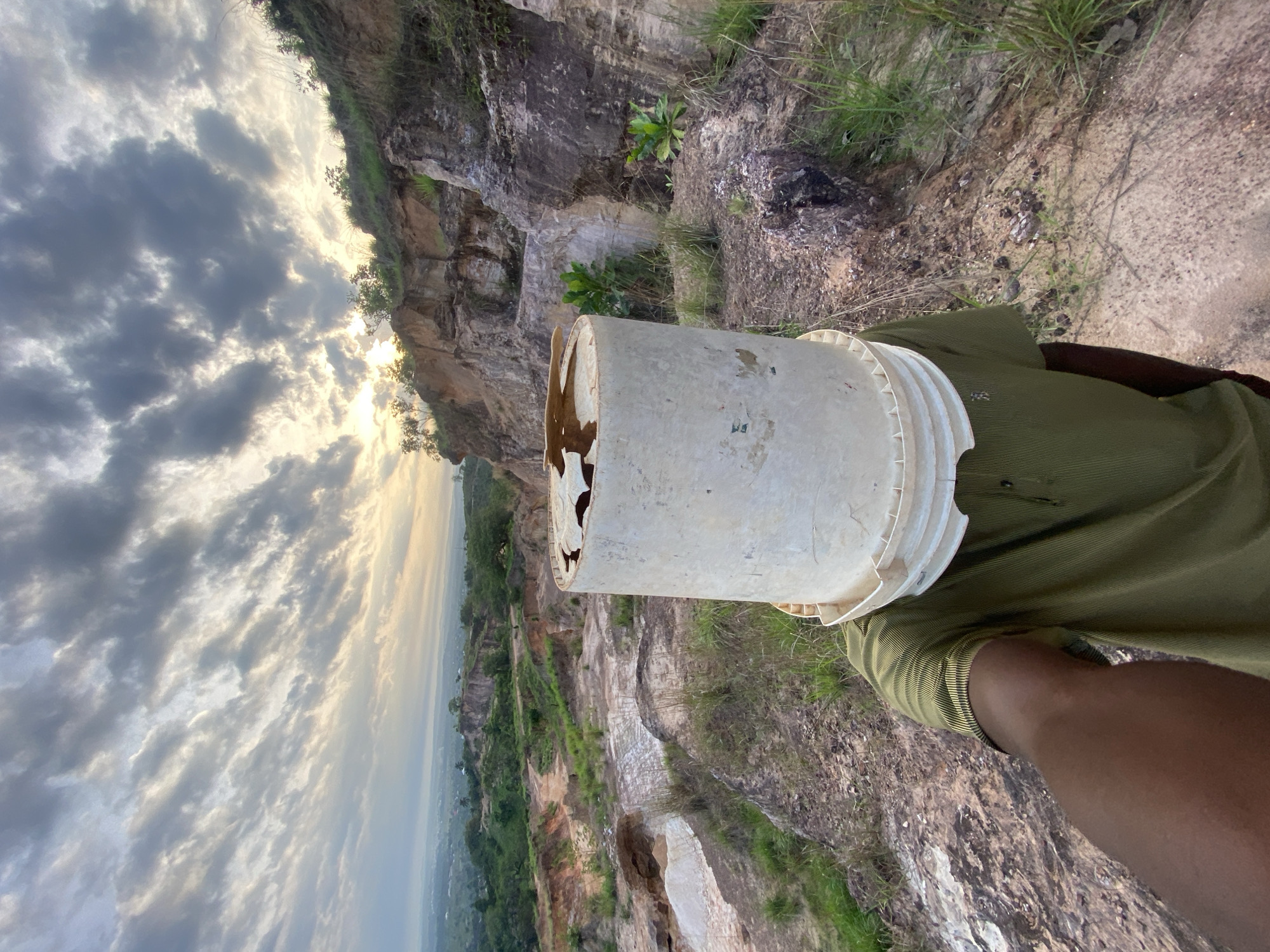 The image, oriented vertically, depicts a person standing on a rocky hillside or quarry in Ufueseni, Nigeria. The central subject is an individual whose head is obscured by a worn, white plastic bucket. The bucket shows signs of age and has a distinct damaged or torn section on one side. The person is wearing a short-sleeved, olive-green polo shirt, and their dark-skinned arm is visible below the bucket.
The background reveals a rugged, natural landscape dominated by large, exposed rock formations and cliffs, characteristic of a quarry or mountainous terrain. Patches of green grass and small plants are interspersed among the rocks. Beyond the immediate foreground, a vast expanse stretches out to the horizon, showcasing a valley or distant settlement under a dramatic sky. The sky is largely covered by white and grey clouds, with the sun partially breaking through, creating a soft, diffuse light that suggests either early morning or late afternoon. No specific activity is clearly depicted beyond the person posing with the bucket on their head, possibly for an artistic or humorous effect. There is no visible text in the image.