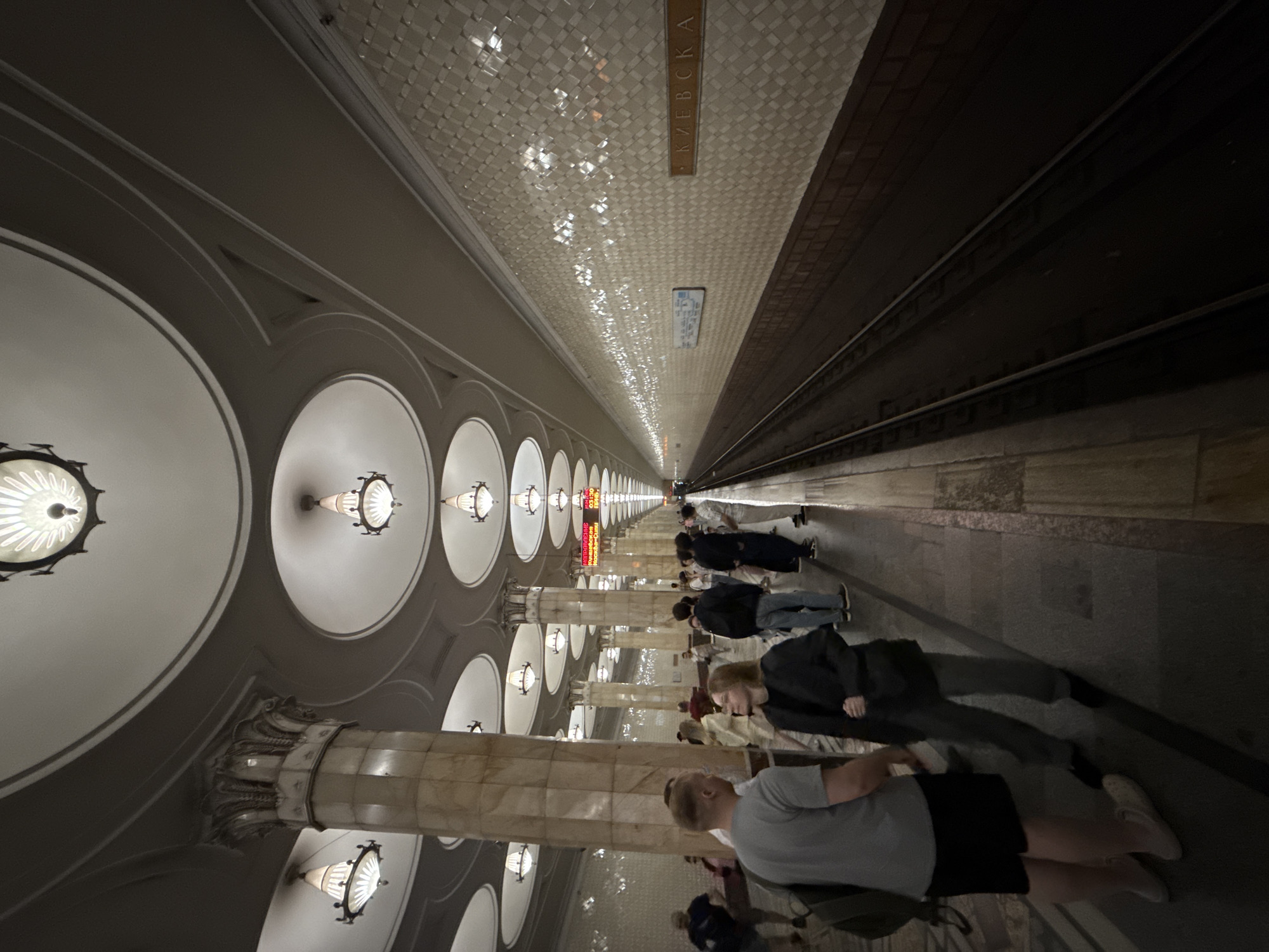 The image depicts the platform of Kiyevskaya metro station in Moscow, Russia. The station's architecture features a high, arched ceiling with ornate lighting fixtures and decorative circular recessed areas. Marble columns, also ornately carved at the top, line the platform. The walls are tiled with a distinctive pattern of small, light-colored squares. A train track runs along the right side of the image, with a train partially visible in the distance. Several people are present on the platform, standing and walking. In the foreground, a man and a woman are facing each other, engaged in conversation. Other individuals are scattered throughout the platform, some appearing to wait for a train. The overall impression is of a grand and well-maintained metro station during a time of moderate passenger activity.