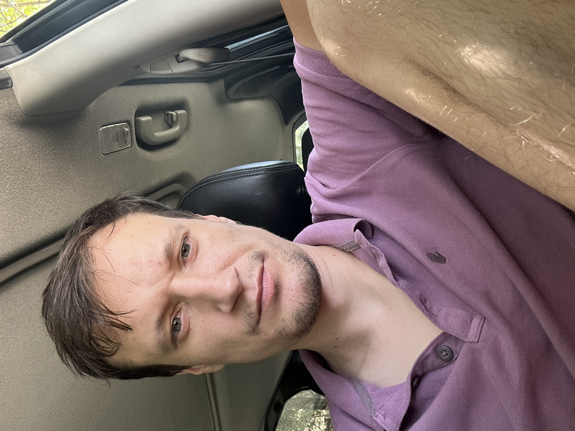 A man is taking a selfie in a car. He is wearing a purple polo shirt. The interior of the car is visible, including the headrest of the passenger seat and the roof lining. The man's right arm and leg are also visible. The image appears to be taken during the day, with natural light coming in from the windows. The location is not discernible from the image itself.