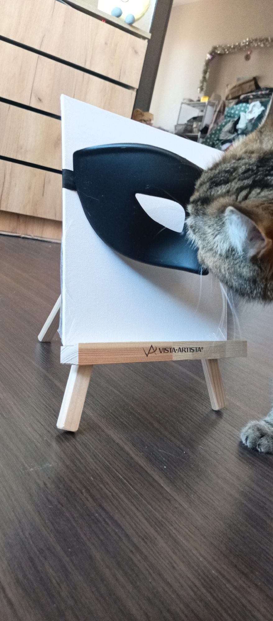A close-up shot shows a cat's head and paw near a small easel holding a blank white canvas with a black mask affixed to it. The easel, labeled "VISTA ARTISTA," stands on a wood-grain floor. The background includes a wooden cabinet with horizontal panels and a cluttered area with tinsel. The location is Unknown, Unknown.