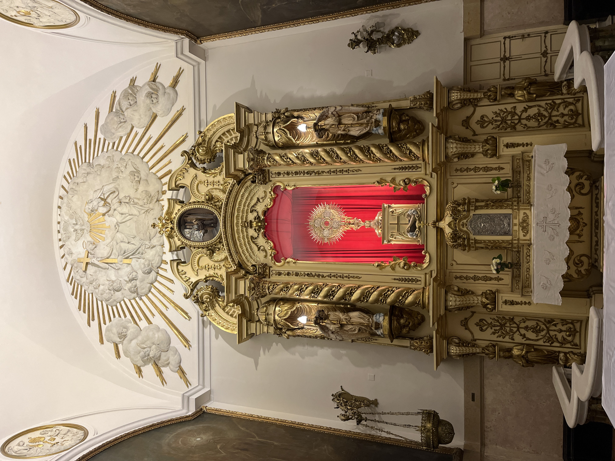 The image displays the richly decorated interior of a church or chapel, likely centered on an altar or reredos, oriented vertically with the altar at the bottom. The dominant features are elaborate gold leaf, detailed carvings, and religious iconography, characteristic of Baroque or Rococo styles.
At the center is a grand, multi-tiered reredos, primarily gilded in gold with creamy white accents. It features intricate scrollwork, columns, and decorative moldings. Within a prominent, arched niche in the middle of the reredos, a golden monstrance with a radiant sunburst design is illuminated against a deep red fabric backdrop. Below this central display, the altar table is covered with a white cloth adorned with embroidered crosses and floral patterns. Small flower arrangements are placed on ledges of the reredos and the altar.
Flanking the central monstrance are two large, ornate golden statues of religious figures, positioned within their own niches or frameworks on either side of the reredos. Above the reredos, a magnificent stucco or plaster relief extends across the vaulted ceiling. This relief depicts numerous cherubic figures and clouds, from which golden rays of light emanate outwards. A prominent golden cross is embedded within this celestial scene, suggesting a divine or heavenly depiction.
The walls are painted a light, neutral color, contrasting with the extensive gilding. Wall-mounted sconces with multiple lights are visible on the side walls, providing additional illumination. On the right, two white chairs are partially visible. A dark painting or panel can be seen on the lower wall to the left. The scene is static, depicting a space arranged for worship or adoration, with no active event taking place. The artificial lighting suggests the time of day is not a factor, or it's an indoor shot. No visible text is present in the image.