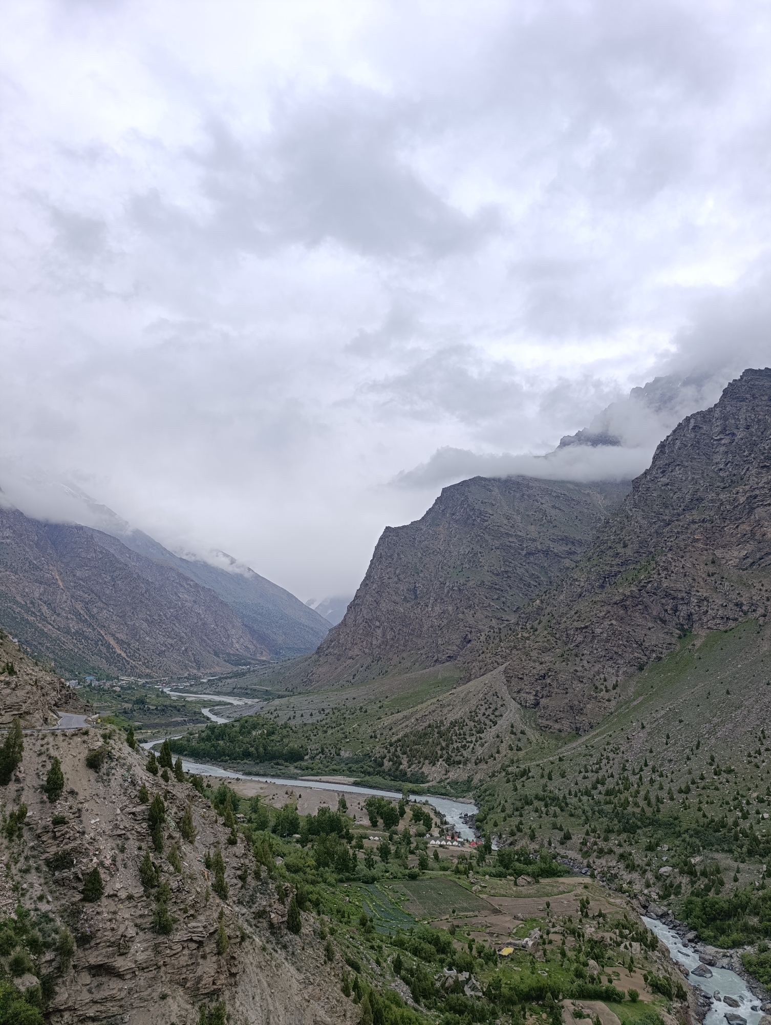 The image shows a mountainous valley with a river flowing through it. The sky is overcast with clouds. The slopes of the mountains are covered with sparse vegetation and rocky outcrops. In the distance, a small village is visible nestled in the valley. The village appears to have a cluster of white tents and some low-lying buildings. A road winds along the side of the valley. The image appears to be taken from a high vantage point, looking down into the valley. The location is specified as Gemur, India.