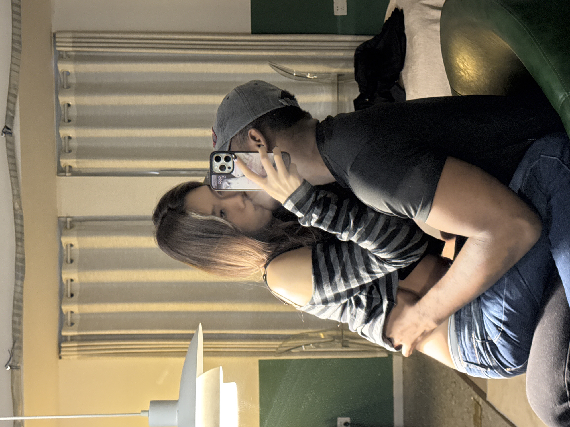 An indoor mirror selfie captures two individuals in a close embrace. The scene takes place within a private room, the specific city of Liangjiang, China is not visually verifiable from the image.
**People:**
A female individual, appearing East Asian with long, dark hair, wears a grey and black striped, off-the-shoulder long-sleeved top and blue denim jeans. A light-colored bra is visible beneath her top. She is seated on the lap of a male individual, leaning in towards him. Her right hand holds a dark-colored smartphone with a triple-lens camera system, positioning it to obscure her mouth and parts of the male's face, indicating she is taking the photograph. Her left arm is around the male's neck. Her fingernails are painted dark.
A male individual, appearing to be a person of color, wears a dark grey baseball cap reversed, a dark short-sleeved t-shirt, and blue denim jeans. He is seated on a green chair. His head is turned towards the female, and his left arm is around her waist. A small stud earring is visible in his left ear.
**Actions and Interactions:**
The female is taking a mirror selfie while seated on the male's lap. Both individuals are in an intimate embrace, with the female's body pressed against the male's. The female's head is positioned as if she is kissing the male, although the smartphone obstructs a direct view of their lips.
**Objects:**
The smartphone held by the female features a transparent or decorative case. A multi-tiered, light grey pendant lamp hangs from the ceiling on the left, illuminated. The male is seated on a dark green, possibly leather or vinyl, chair. In the background, light-colored, floor-length curtains with eyelet rings hang against a lighter-toned upper wall. A light-colored surface, possibly a bed or table draped with a cloth, is visible behind the male's right shoulder, with a dark item of clothing resting upon it. A faint reflection of a clear plastic chair or stool is visible behind the male's head. White electrical outlets are visible on the lower green wall section on both the left and right sides of the frame.
**Environment:**
The room features a lower wall section painted dark green and an upper section painted a lighter, possibly yellow or beige, color. The floor is light-colored and somewhat reflective. The primary illumination appears to be from artificial light sources within the room, including the visible pendant lamp, casting a warm tone.