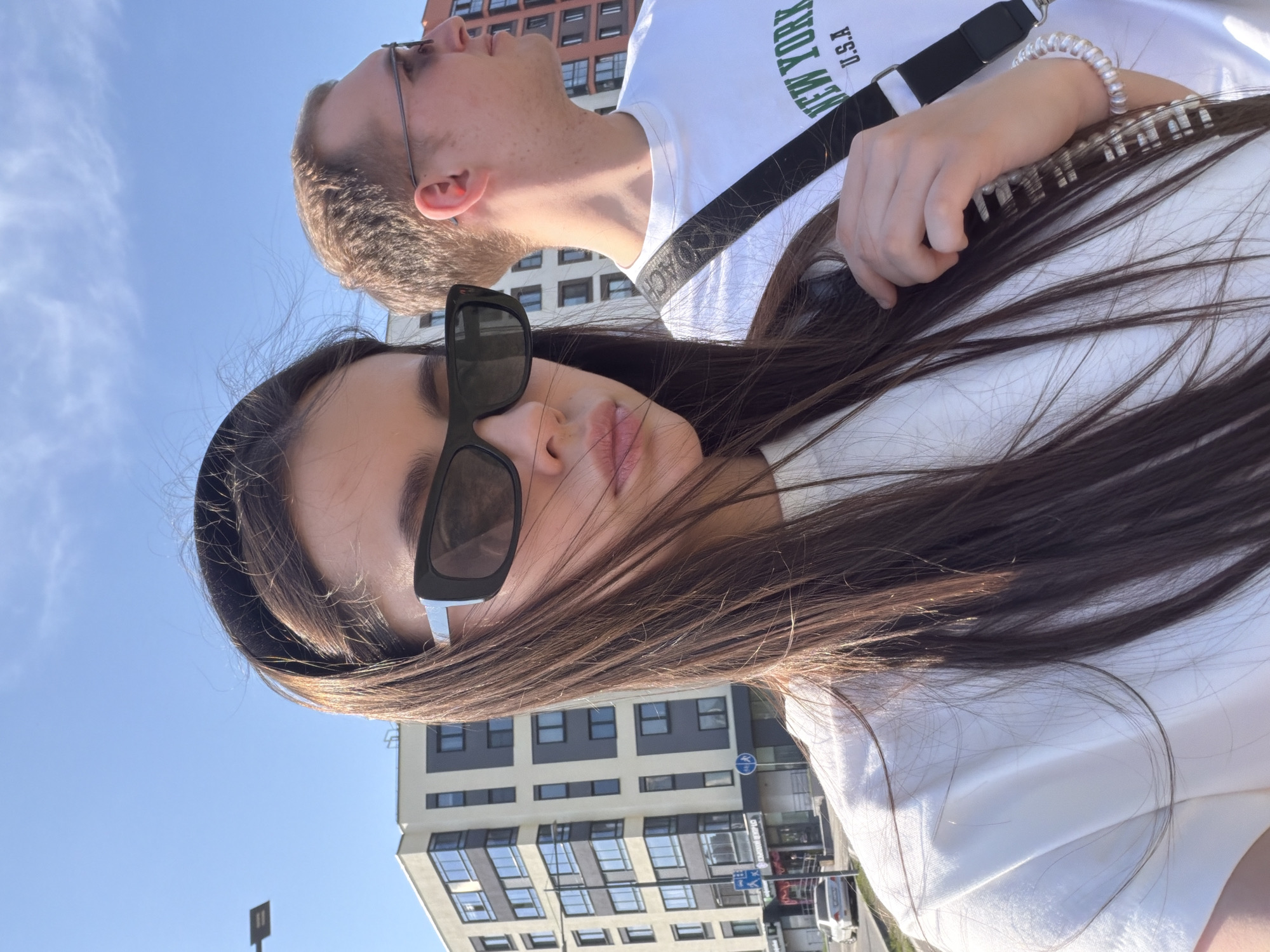 A selfie shot from a low angle shows a young woman with long brown hair and large, dark sunglasses in the foreground. A young man with glasses and short brown hair is slightly out of focus in the background. Both individuals are wearing white t-shirts. The woman has a white bracelet on her right wrist and appears to be holding a bag strap. Buildings are visible in the background, including one with many windows, indicative of a modern apartment complex. The setting is outdoors on a sunny day with a clear blue sky. Based on the distinct architectural features visible in the background, the location appears to be in Izhevsk, Russia, specifically near the "Flame Tower Izhevsk" apartment building, with the adjacent street and intersection.