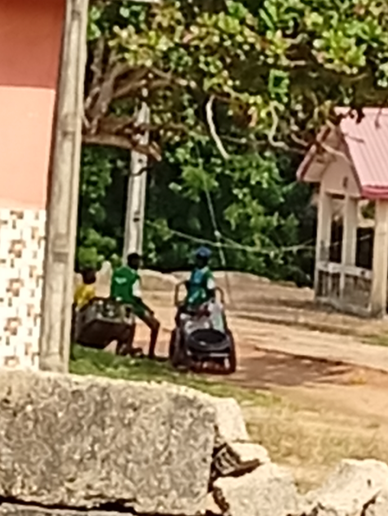 A group of young people are interacting outdoors in a rural or suburban setting. Two individuals are standing and appear to be engaged in an activity with a large, dark, rectangular object, possibly a container or piece of equipment. One of these individuals is wearing a green shirt and shorts. A third person is seated in a wheelchair or similar apparatus, wearing a blue cap and a green shirt. In the background, there are trees with green foliage and a light-colored building with a pink roof. The ground appears to be a dirt road or path, with patches of grass. A stone structure is visible in the foreground. The lighting suggests it is daytime.