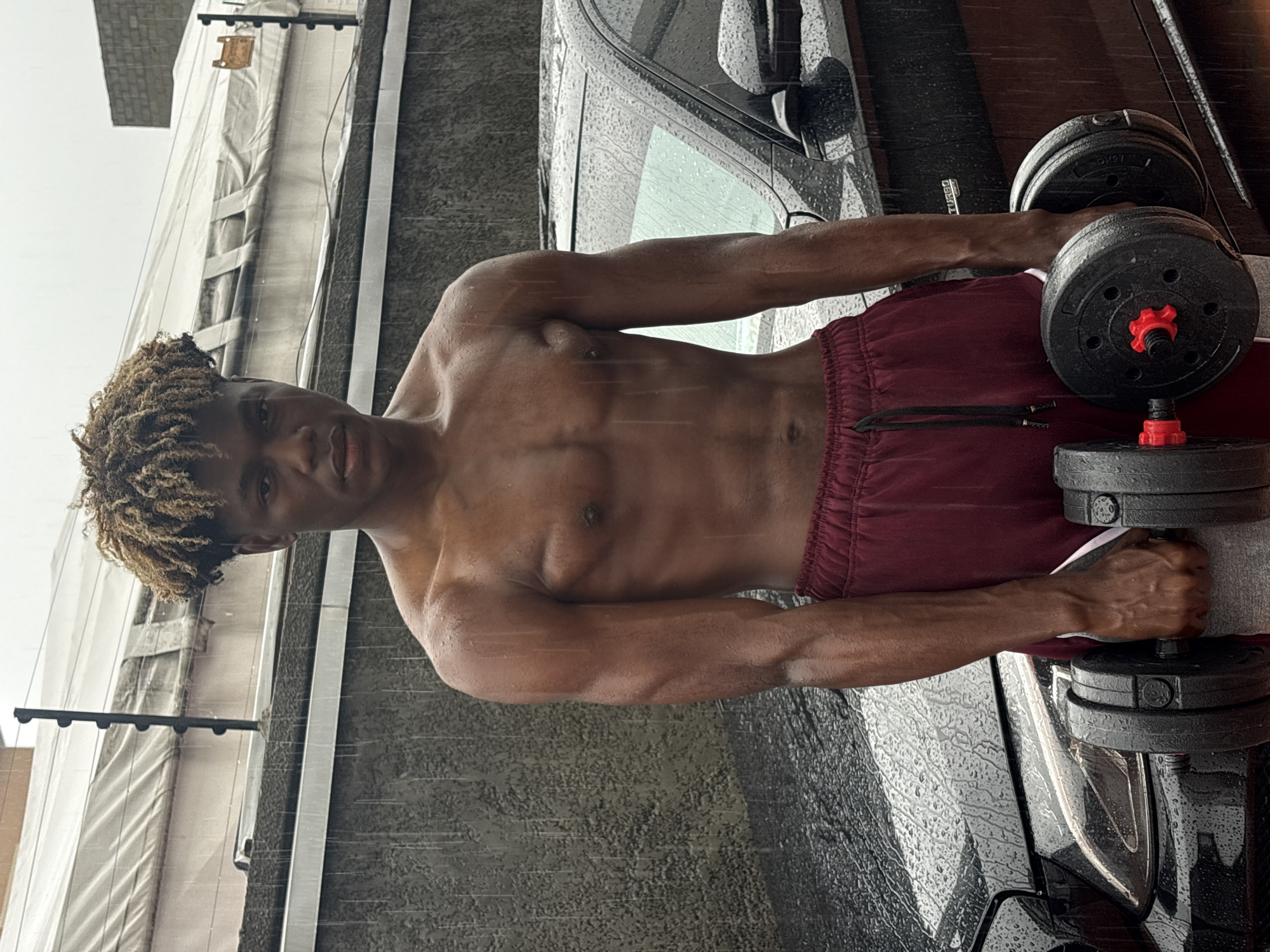 The image shows a shirtless man with short, dyed-blonde hair standing in what appears to be the Igboefon area of Nigeria. He is holding two dumbbells with black weights and red adjustment knobs in front of his body, one in each hand. He is wearing red shorts. Rain is visible on the man's skin, a black car, and the textured wall behind him. The background includes a building with white siding.