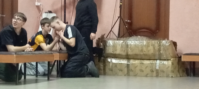 The image depicts an indoor scene in Красный Маяк, Russia, featuring four males. In the foreground, two young adult males are engaged in arm wrestling across a dark brown rectangular table. The male on the left of the arm wrestling pair, wearing a black and yellow shirt, kneels with his right elbow on the table. The male on the right of the arm wrestling pair, wearing a dark shirt with white stripes on the shoulder, also kneels with his left elbow on the table, appearing focused. To the left of the arm wrestling setup, a third young adult male with glasses, wearing a dark short-sleeved shirt, sits on a bench, observing the activity with an open mouth. In the mid-background, a fourth male, dressed in a dark hoodie and dark pants, stands partially visible behind the arm wrestlers, facing right.
The room features light pink/peach colored walls, with a white electrical outlet or switch visible on the left wall. The floor is light-colored. A black tripod stand is visible in the background, to the right of the standing male. Against the right wall, two large stacks of cardboard boxes wrapped in clear plastic or tape are present. A portion of a light brown bench with a dark cushion is visible on the far right. A dark wooden door or paneling is seen on the right side of the background.