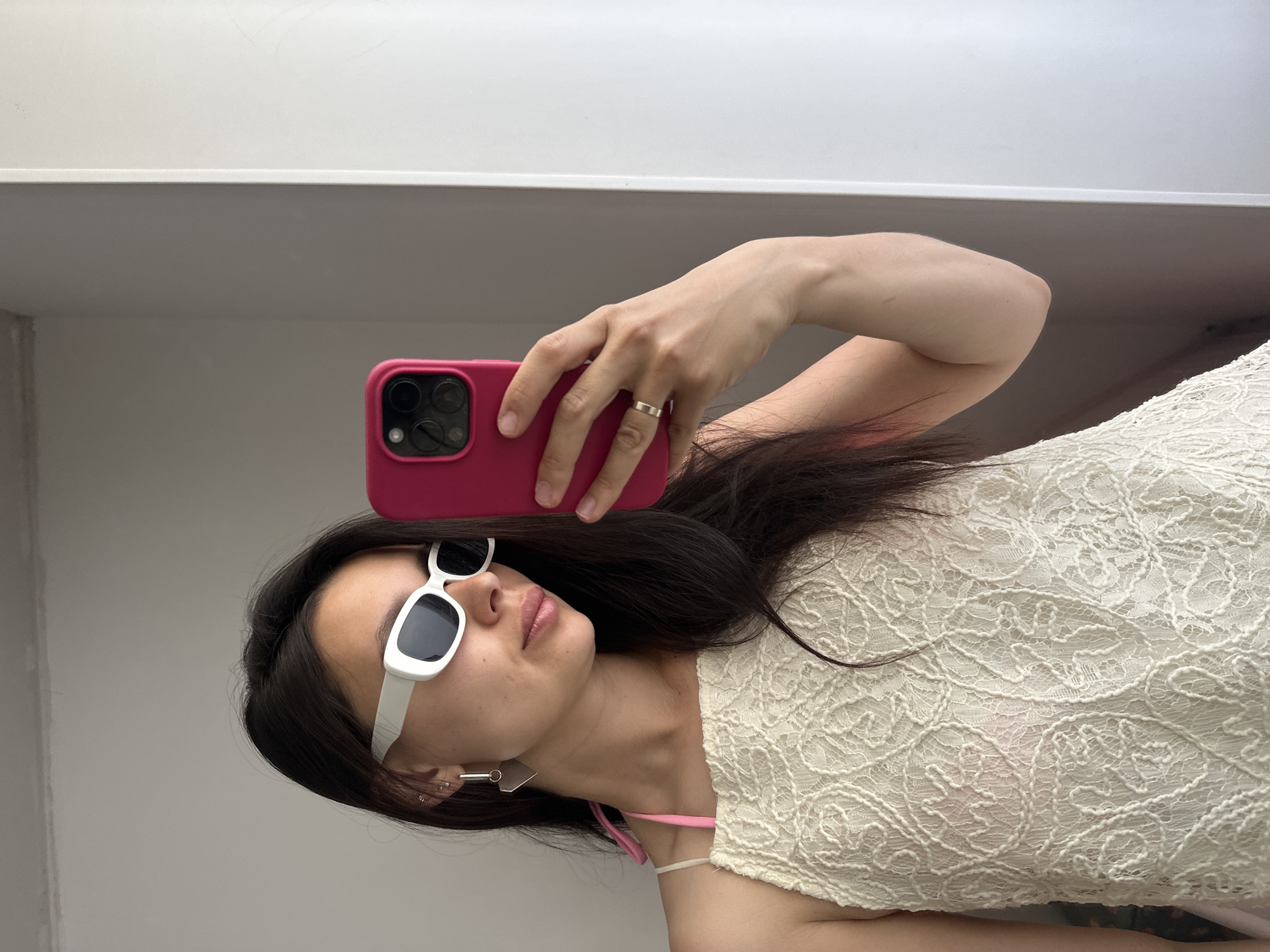 A young woman is taking a selfie in front of a neutral white background. She is wearing white-framed sunglasses and an off-white, lace-patterned top. She is holding a pink phone case in her right hand, which has a silver ring on the ring finger. Her hair is long and dark. There is a pink strap visible under her top. Her left ear has multiple piercings and a geometric earring. It is impossible to determine the location in the city of Chaikovsky, Russia from this image.