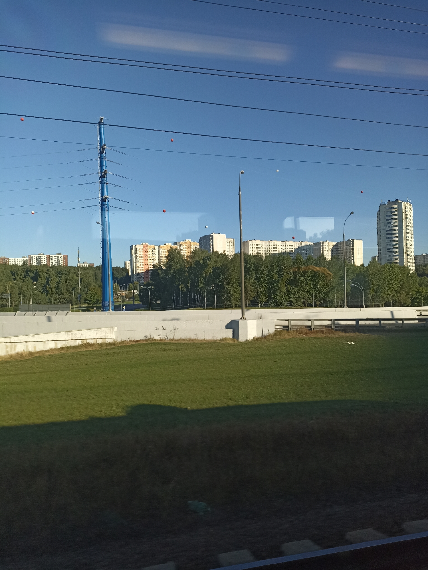 The image shows a view of a cityscape, likely in Moscow, Russia, taken from a moving vehicle. In the foreground, a green grassy area is visible, with a dark shadow cast across it, suggesting the sun is low in the sky. Behind the grass, a white concrete barrier runs horizontally across the frame. Beyond the barrier, a dense line of trees forms a green buffer.
Further in the background, several multi-story apartment buildings are visible, with a mix of colors and architectural styles. Some of these buildings are tall and modern, while others appear older. A prominent blue electricity pylon stands out against the sky, with several power lines stretching horizontally across the upper portion of the image.
Several small, orange-red spherical objects are suspended in the sky, seemingly attached to or hanging from the power lines. These could be decorative elements or warning markers. A few white objects, possibly balloons or more markers, are also present in the sky. Streetlights are visible along the road behind the concrete barrier. The sky is a clear blue with faint wisps of clouds. The overall impression is of a suburban or urban residential area on a clear day.