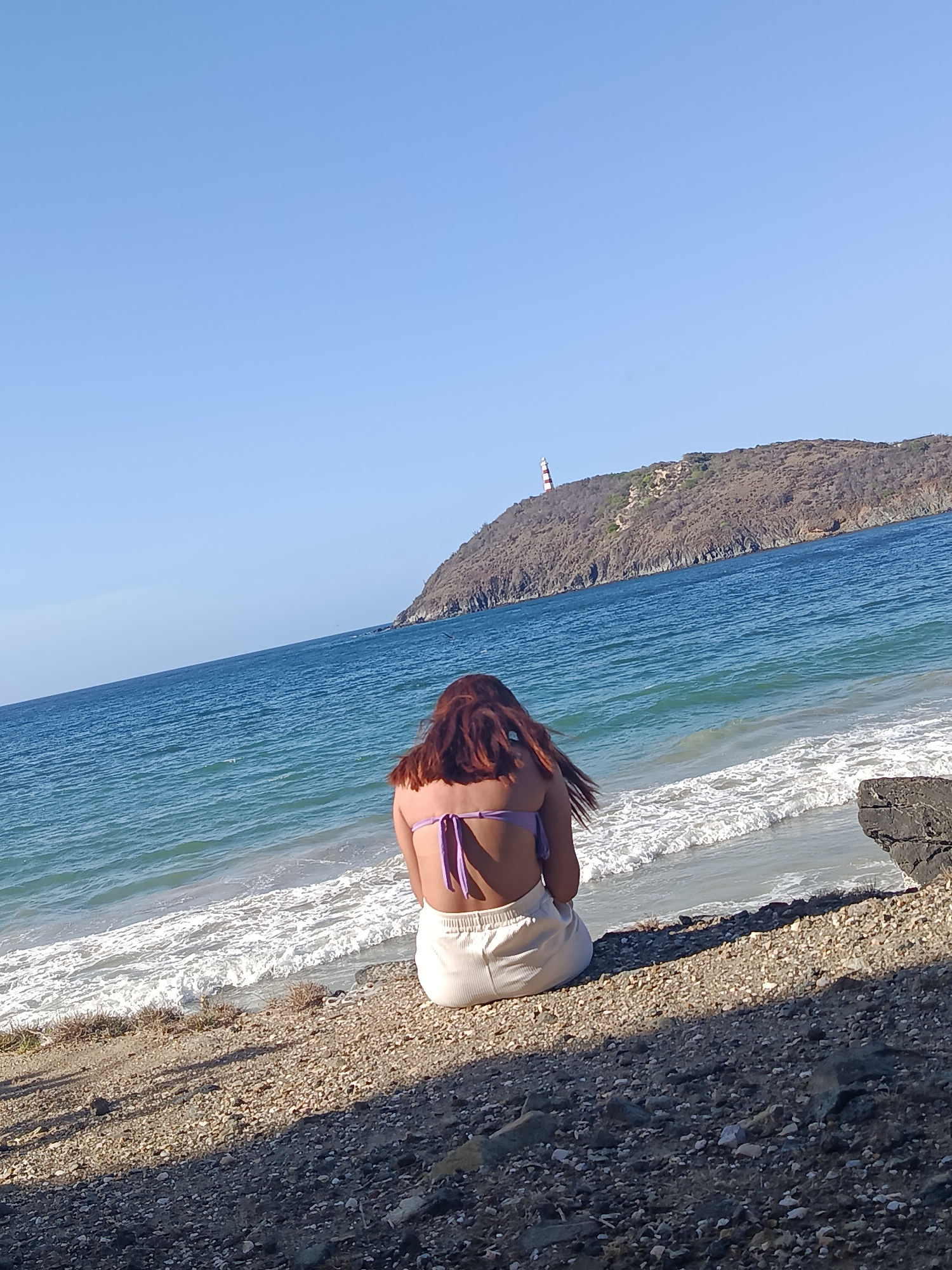 A woman with long, auburn hair sits facing the ocean on a pebble beach. She is wearing a purple bikini top and cream-colored shorts. The ocean is a vibrant blue with white waves breaking on the shore. In the background, a lighthouse with red and white stripes stands on a hill overlooking the water. The sky is clear and blue.