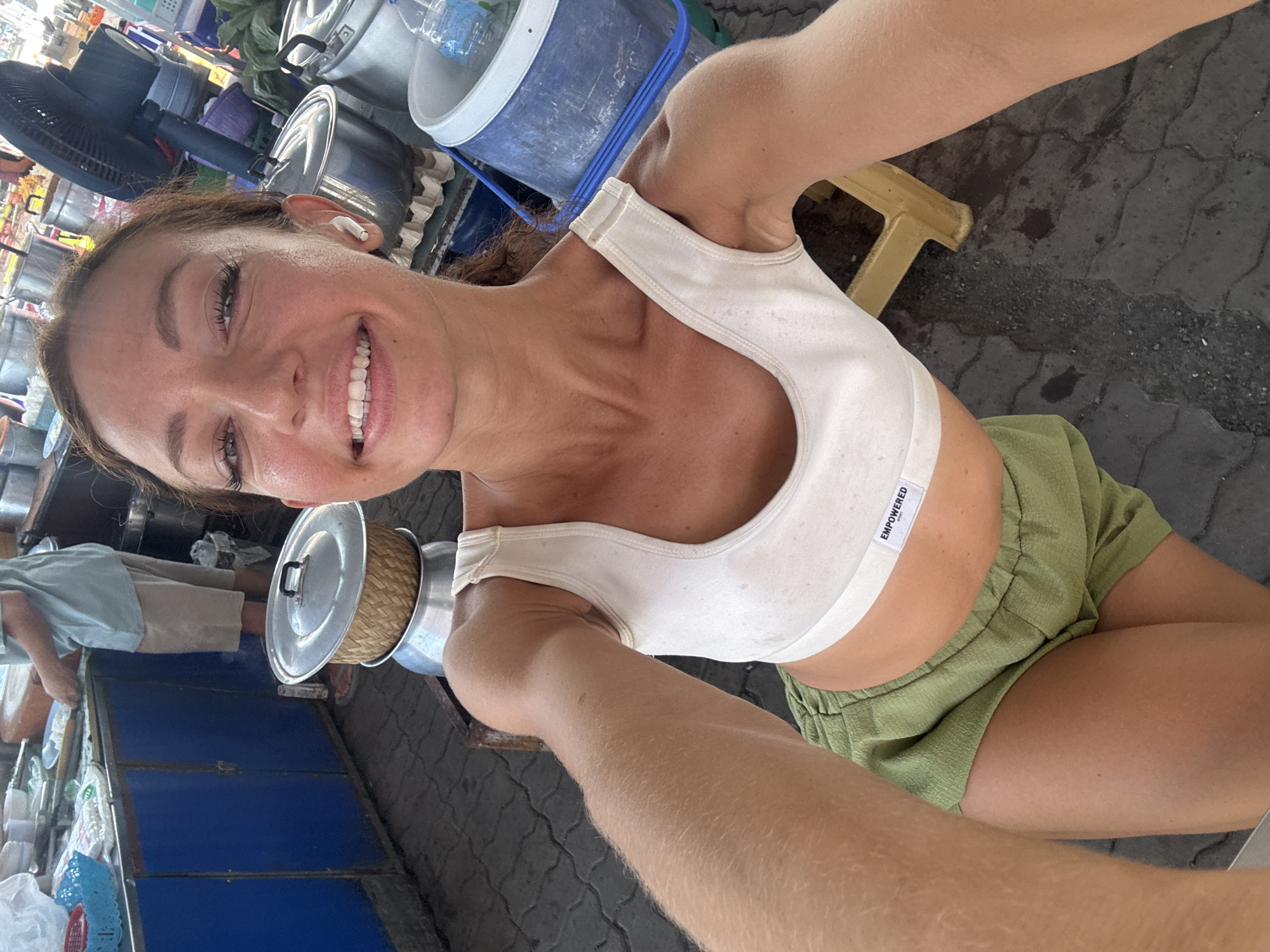 A woman with brown hair, wearing a white sports bra and green shorts, is holding a silver pot with a lid in her left hand, possibly at a street food vendor. The woman is smiling at the camera, and has an AirPod in her left ear. Behind her is a street scene with food vendors, including metal pots and a blue vendor cart. The location is likely in the city of บ้านบางรักษ์, Thailand. The ground is paved and there is a small yellow stool visible.