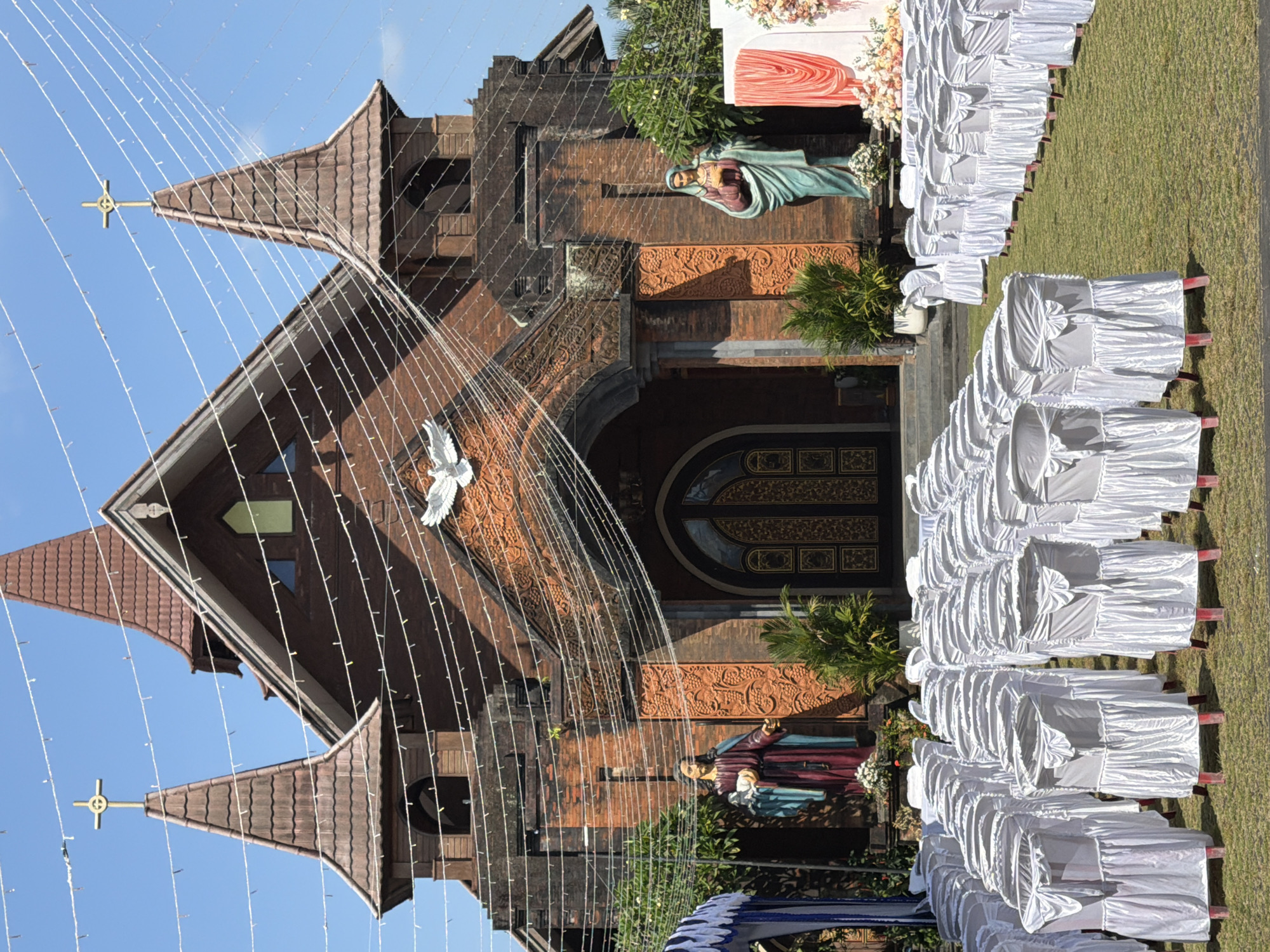 The image depicts the exterior of a church in North Kuta, Indonesia, with rows of white-covered chairs set up on a grassy lawn in front of it, suggesting an outdoor event or ceremony. The church is constructed of red brick and features two prominent spires topped with crosses. Decorative white string lights are strung across the facade of the building. Two statues of religious figures stand on either side of the entrance: a male figure to the left, and a female figure to the right. The entrance itself is an ornate double door. A white dove is depicted in relief above the entrance. The sky is clear and blue. The lighting suggests it is daytime.