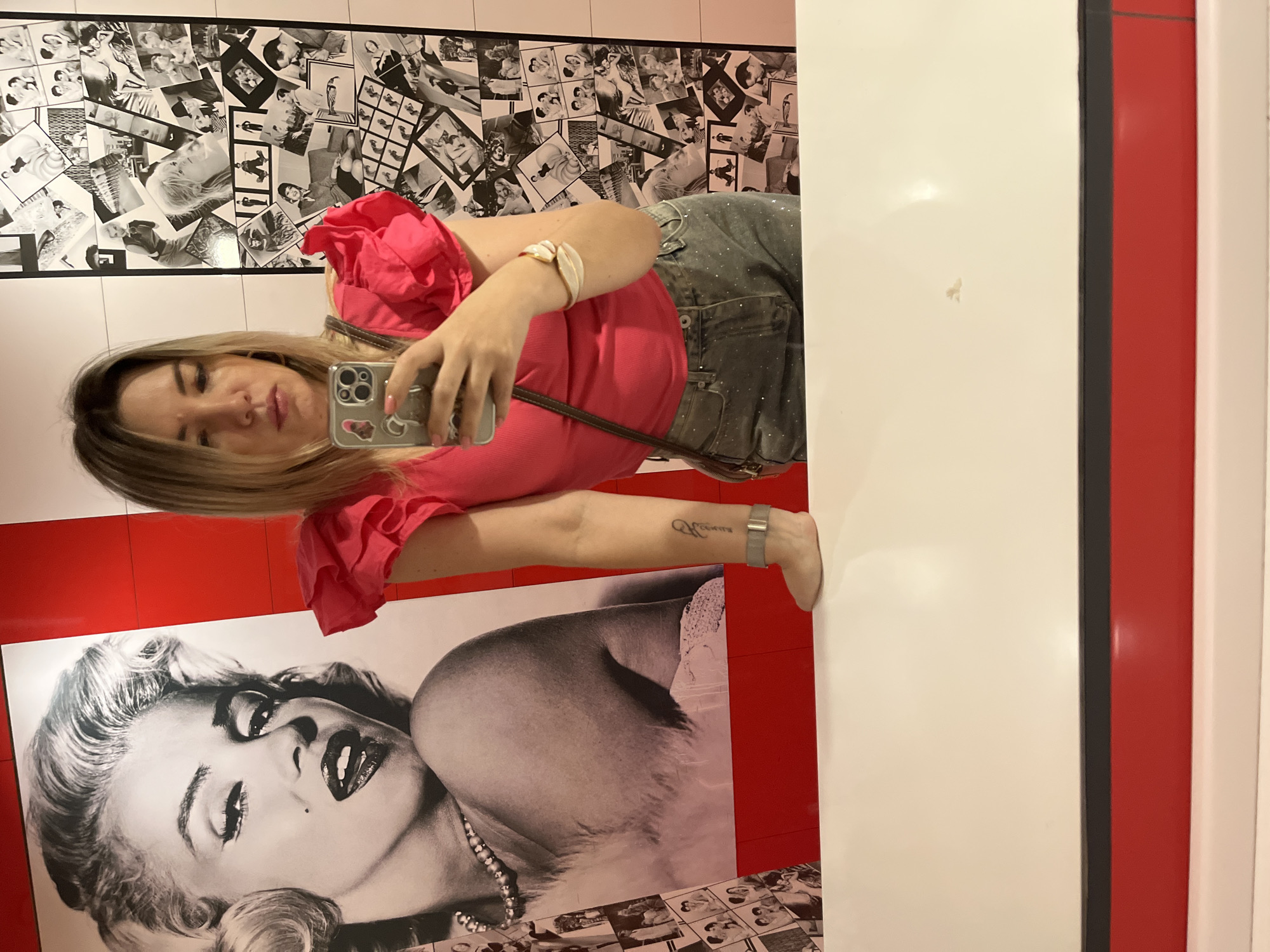 A female subject is captured in a mirror selfie within an indoor setting in Sochi, Russia. The subject has shoulder-length blonde hair and is wearing a hot pink top with voluminous ruffled sleeves and dark grey denim jeans embellished with small sparkles. She holds a silver-colored smartphone with a clear, sticker-adorned case in her right hand, positioned to capture her reflection. A dark brown shoulder bag strap is visible across her torso. On her left wrist, she wears a light-colored, leaf-shaped bracelet, and a small, vertical script tattoo is present on her left forearm. Her left arm rests on a white counter that occupies the lower portion of the frame. Her right foot is also visible on this counter surface.
The background behind the subject features two distinct decorative wall sections. To the left, a large black and white poster depicts Marilyn Monroe from the chest up, wearing a pearl necklace, with an open-mouthed expression. The wall area immediately surrounding this poster and behind the subject's left arm is solid red. To the right of the subject, the wall is covered with a collage of multiple smaller black and white photographs, showcasing various classic film and fashion icons and scenes. This collage is bordered by white wall segments. Below the white counter, a thin dark grey strip precedes a wider, solid red panel. The environment is illuminated by artificial light.