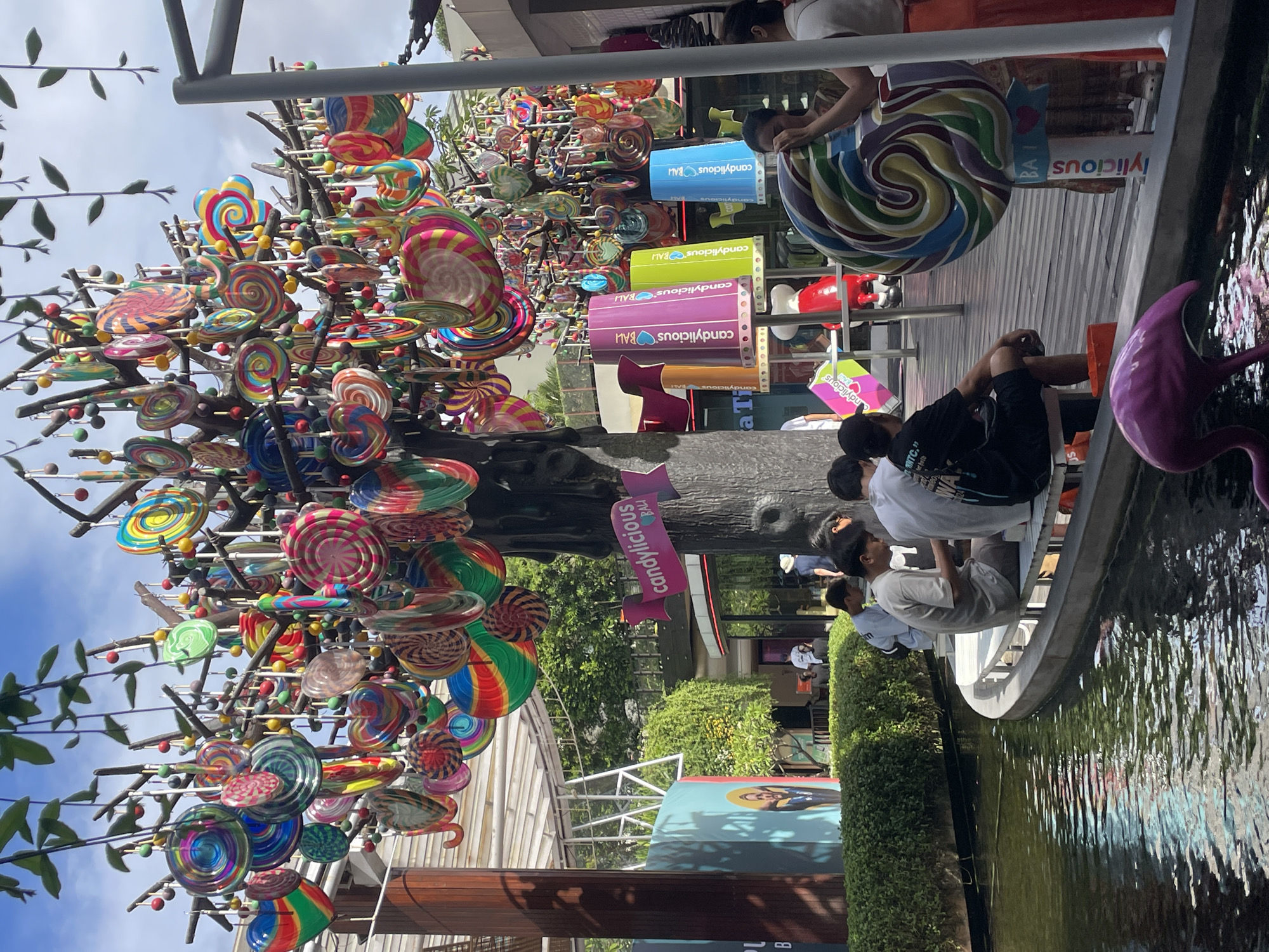 This vibrant outdoor scene captures a candy-themed area, likely a store or attraction, under a bright daytime sky. The central feature is a large, whimsical "candy tree" adorned with numerous colorful, swirl-patterned lollipops and other candy-shaped decorations, creating a playful atmosphere. Many of these lollipop decorations are in various sizes and vibrant hues, including rainbow, red, blue, green, and yellow patterns.
Several people, mostly young adults, are visible. Some are seated on a low ledge beside a body of water, which shows rippling reflections. One person wears a black t-shirt with "NTC." partially visible. Another large, multi-colored swirl lollipop decoration stands prominently on a deck behind the seated individuals, and a pink flamingo statue is positioned near the water's edge.
Various advertising banners and cylindrical displays are strategically placed, repeatedly featuring the brand "Candylicious BALI" in different colors such as blue, lime green, and purple. A pink banner also clearly displays "candylicious." The background includes green hedges, foliage, and parts of buildings, suggesting a leisure or entertainment complex. The clear blue sky indicates sunny weather during the day. The overall impression is one of a cheerful and sweet-themed environment where people are relaxing.