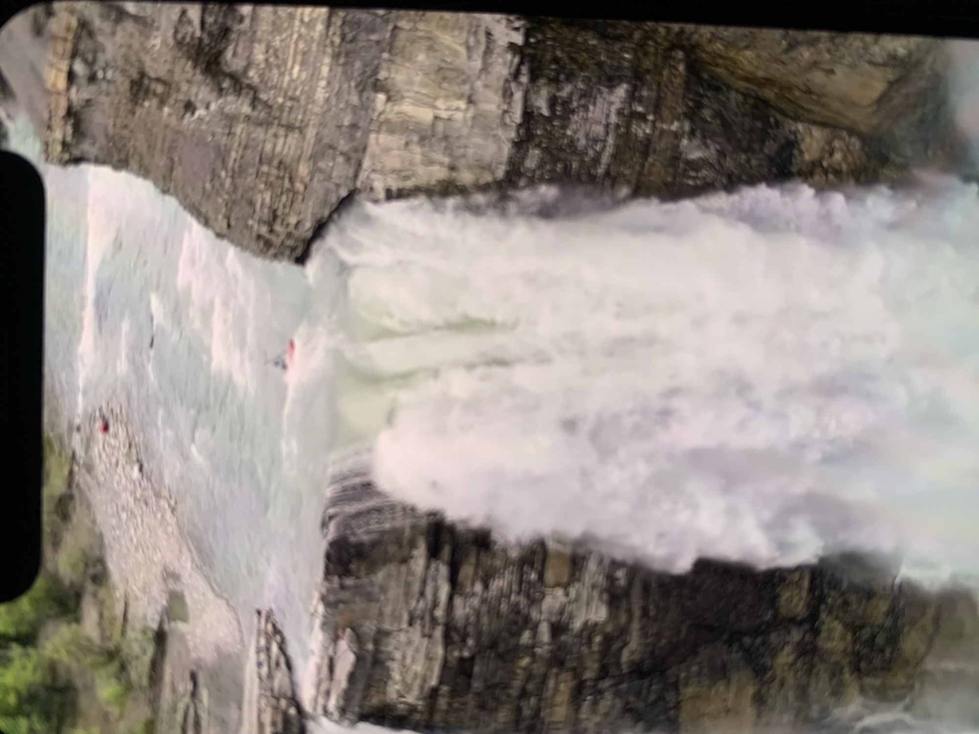 The image shows a wide waterfall cascading over a rocky cliff face. A river flows at the top of the waterfall and continues below. There are at least two red kayaks visible in the river. One kayak is near the top of the waterfall, and another is further upstream. The surrounding environment appears to be natural, with trees visible on the banks of the river in the distance. The image is taken from an elevated perspective, looking down at the waterfall and the kayaks. The location is not Stavropol, Russia, as the geography does not match.
