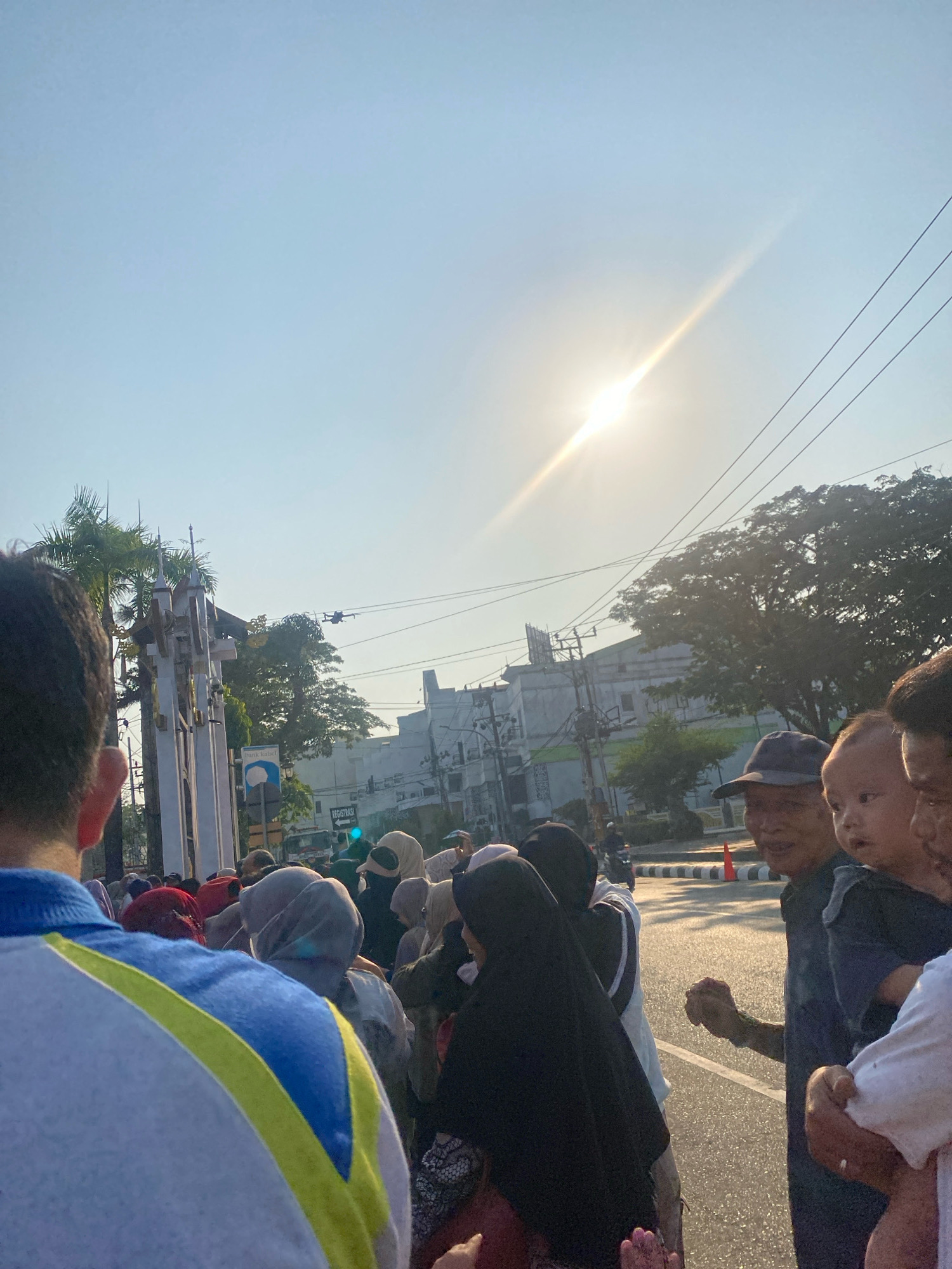 In Banjarmasin, Indonesia, a crowd of people, many wearing headscarves, gathers on a street on a clear, sunny day. The sun is high in the sky, casting a bright glare. In the foreground, the back of a person wearing a blue and yellow shirt is visible. To the right, several individuals are in focus, including an older man wearing a baseball cap, a baby being held by an adult, and another person in a white shirt. In the background, a tall, ornate monument stands to the left, with palm trees and other greenery. Across the street, buildings and utility poles are visible, along with a drone flying overhead. A sign with the word "REGISTRASI" is also present. The atmosphere appears to be peaceful, possibly a religious gathering or public event.