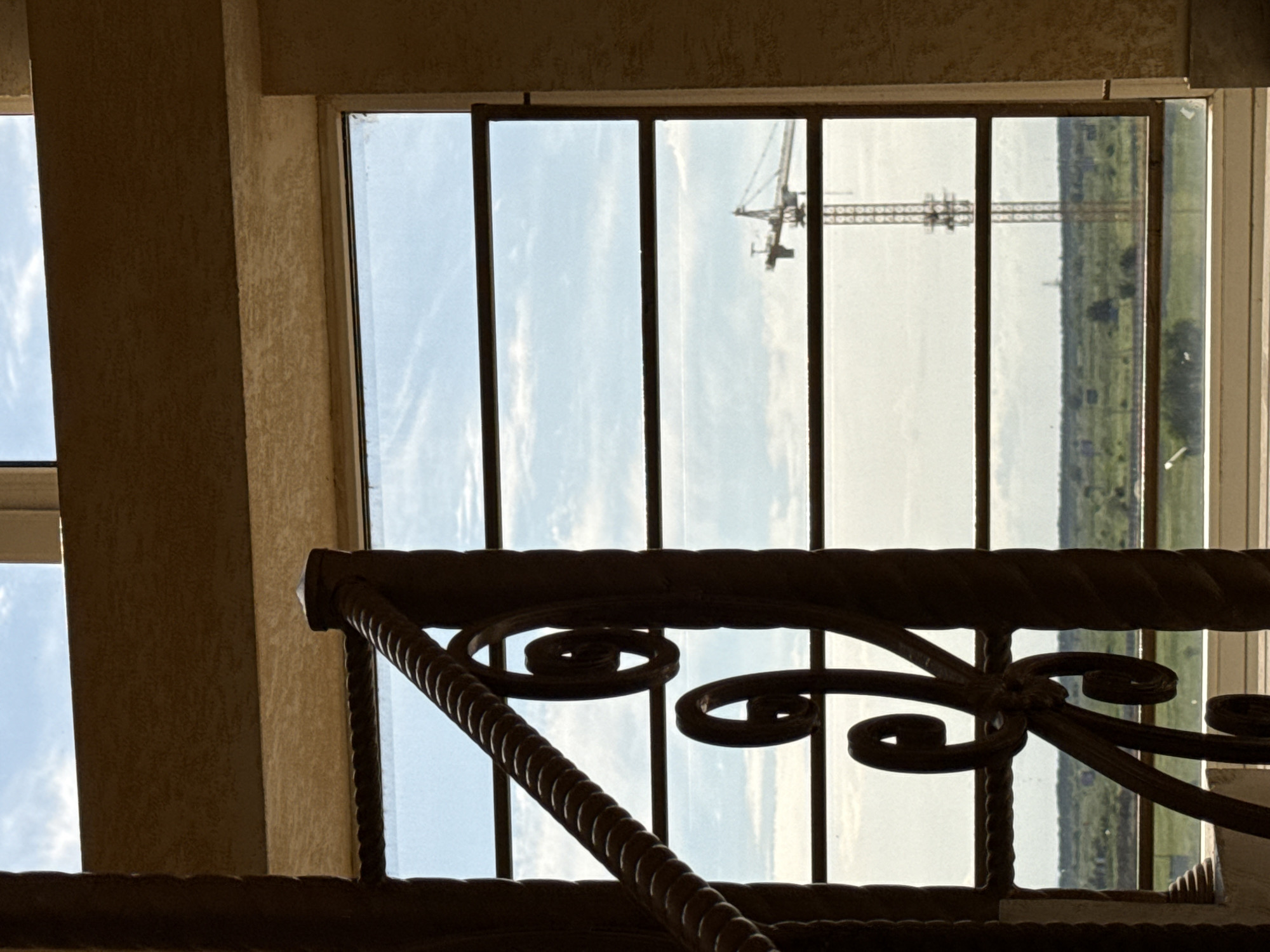 The media file presents an indoor perspective looking out through a multi-paned window. In the foreground, a dark, silhouetted wrought iron railing with decorative scrolls and twisted elements partially obstructs the view. To the left and surrounding the window, sections of textured, light brown building walls or pillars are visible, indicating an interior setting.
Through the window, a bright blue sky with sparse, wispy white clouds dominates the upper half of the outdoor scene. In the mid-ground distance, the distinctive silhouette of a large construction crane, likely a tower crane, is clearly visible against the sky. The lower part of the distant view reveals a flat, green landscape, possibly fields or distant trees, stretching towards the horizon. The light suggests it is daytime, likely a bright morning or afternoon, with clear weather. There is no explicit activity taking place, and no people are visible. No text is discernible in the image.