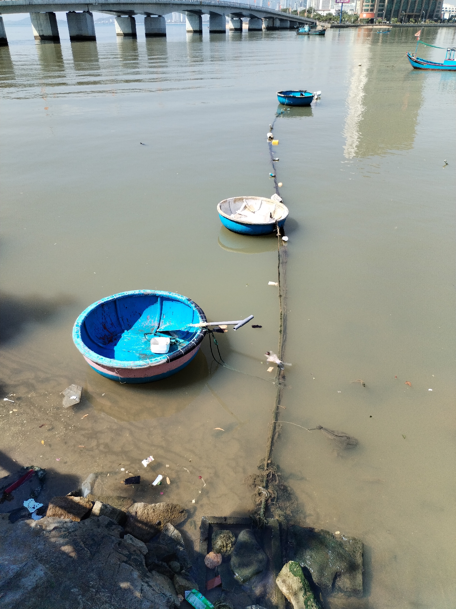 The image shows a polluted waterway in Bac Nha Trang Ward, Vietnam. Three round, blue-rimmed fishing boats are tethered together in shallow, murky water. Debris litters the shoreline, which is composed of rocks and concrete. A bridge is visible in the background, and tall buildings are on the opposite shore. The overall scene suggests environmental degradation.