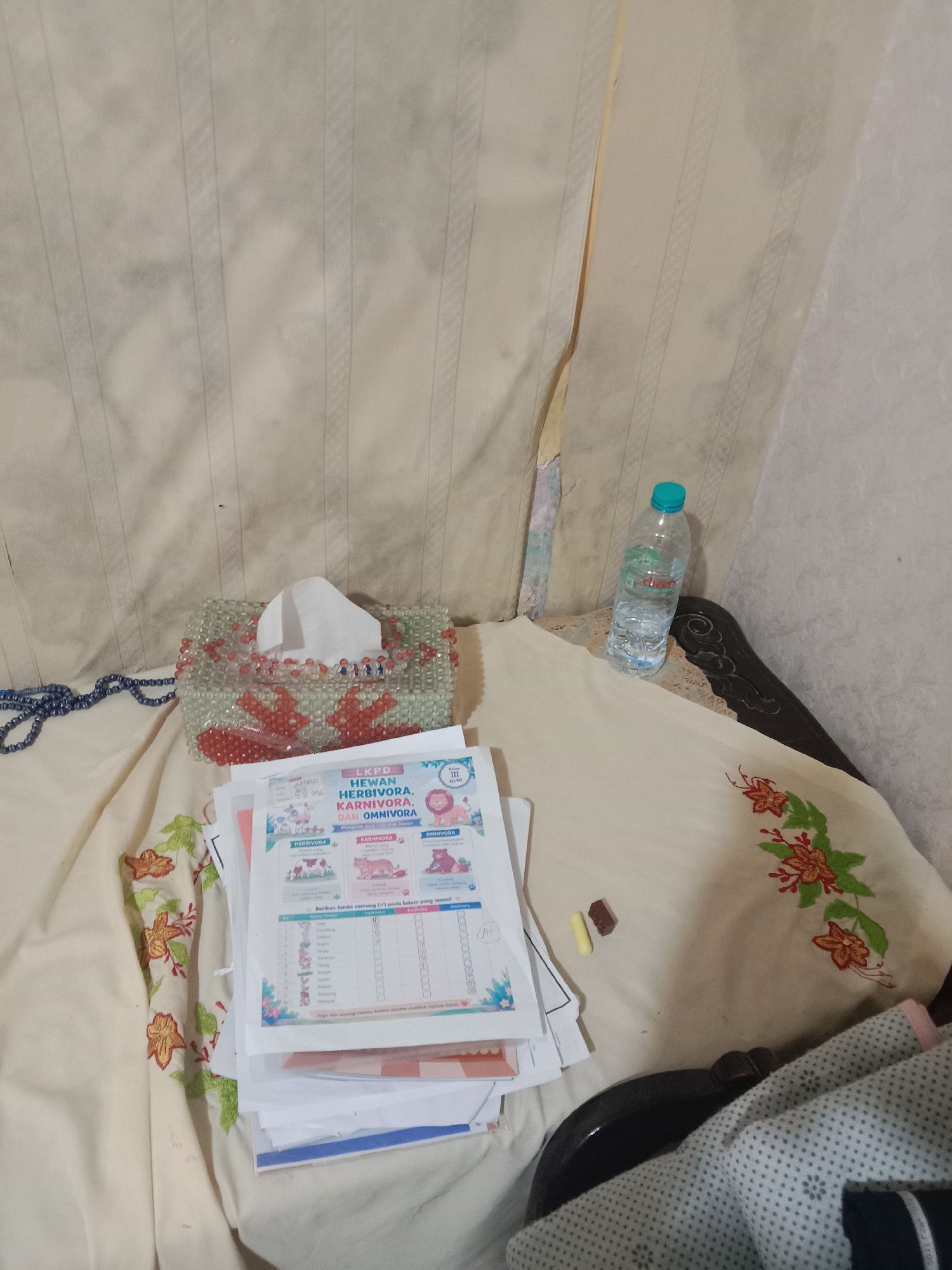 The image shows a close-up of a bed or sofa with various items scattered on it. In the center, a decorative tissue box made of beads is visible. In front of the tissue box, a stack of papers with educational content, possibly for children, is laid out. The papers appear to be about herbivores, carnivores, and omnivores, with illustrations of animals. To the right of the papers, a single bottle of water with a blue cap is standing upright. In the background, a wall with vertical stripes and a torn piece of wallpaper is visible. On the left side of the image, a beaded blue necklace is draped across the fabric. A small yellow and brown object, possibly a piece of candy or a marker, is placed on the fabric near the papers. The overall scene suggests a casual, indoor environment, likely a bedroom or living area. The image is an interior shot and does not provide any contextual clues to determine its location in Madiun, Indonesia.
