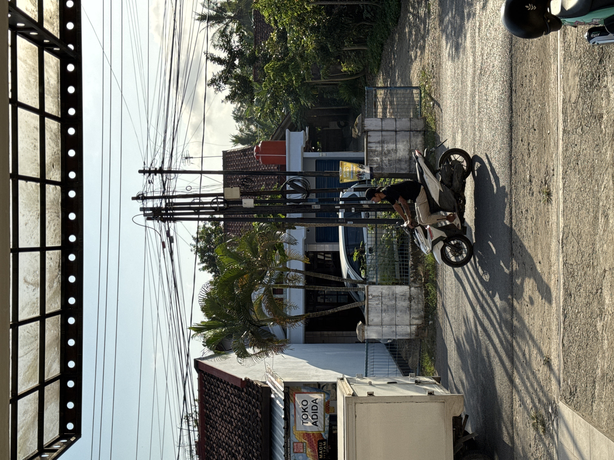 In Kedawung, Indonesia, a man wearing a black t-shirt and tan pants rides a white scooter. He is seen from the side, moving from left to right across the frame. The scooter is casting a long shadow on the asphalt road. In the background, a white truck is parked on the left, and a house with a red tile roof is visible on the left side of the image. There are palm trees and other foliage around the house, and utility poles with many wires crisscross the sky. The scene appears to be a street in a residential or commercial area, possibly with a shop called "TOKO ADIDA" indicated by a sign. The overall atmosphere is bright and sunny, with the light casting distinct shadows.