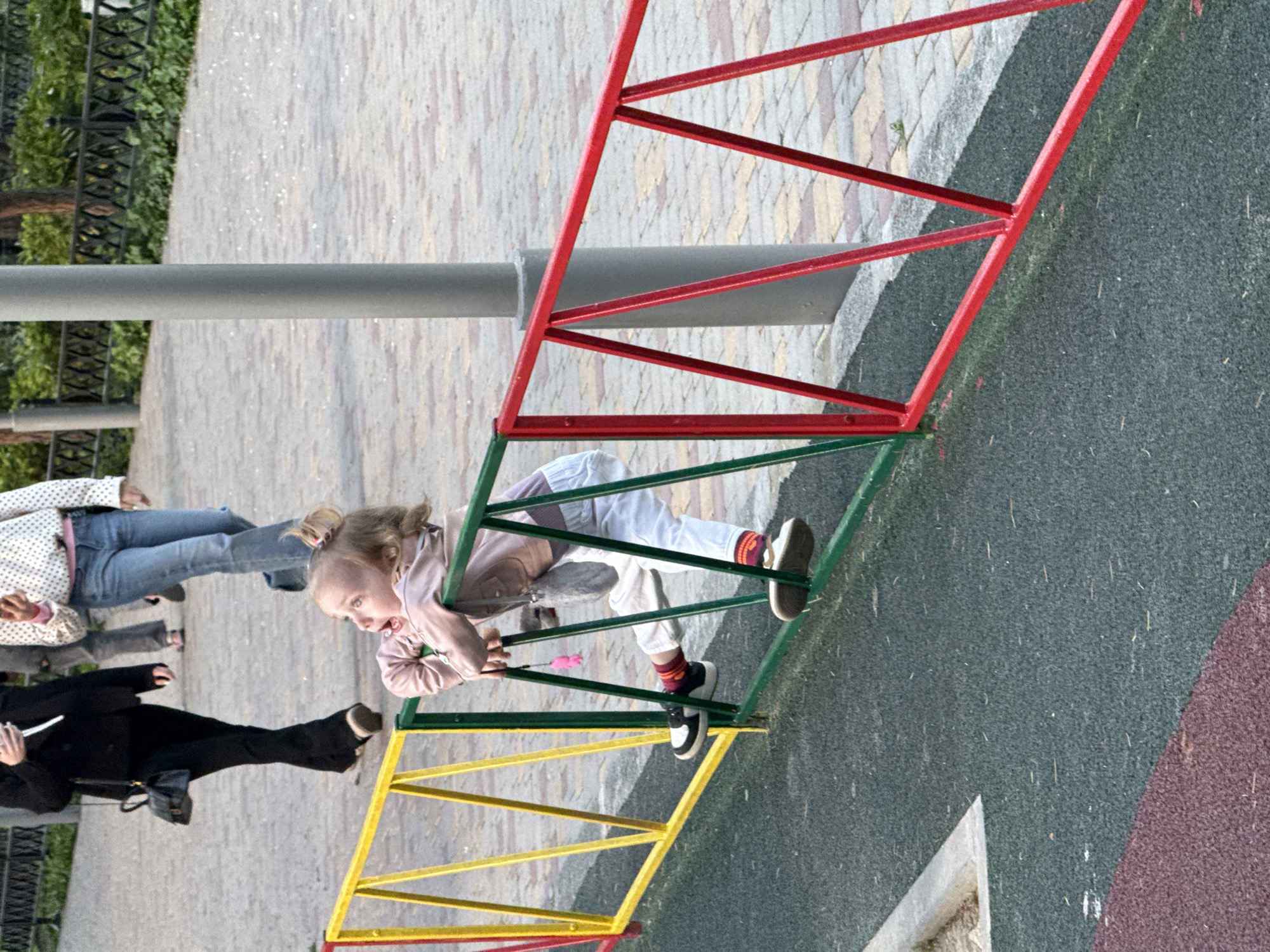 A tilted outdoor scene in Unknown, Unknown, shows a young girl playing on playground equipment. The colorful equipment features red, green, and yellow metal bars in a geometric pattern. The girl, wearing light-colored pants, a pink jacket, and sneakers, is holding onto the bars. There are two people standing on a gray pavement, one in a black suit, and one in jeans and a white shirt with black dots. The ground is a mix of gray playground surface and a brick-like pattern. A metal pole extends horizontally across the image, partially obstructing the view of the playground equipment. Vegetation is visible in the background.