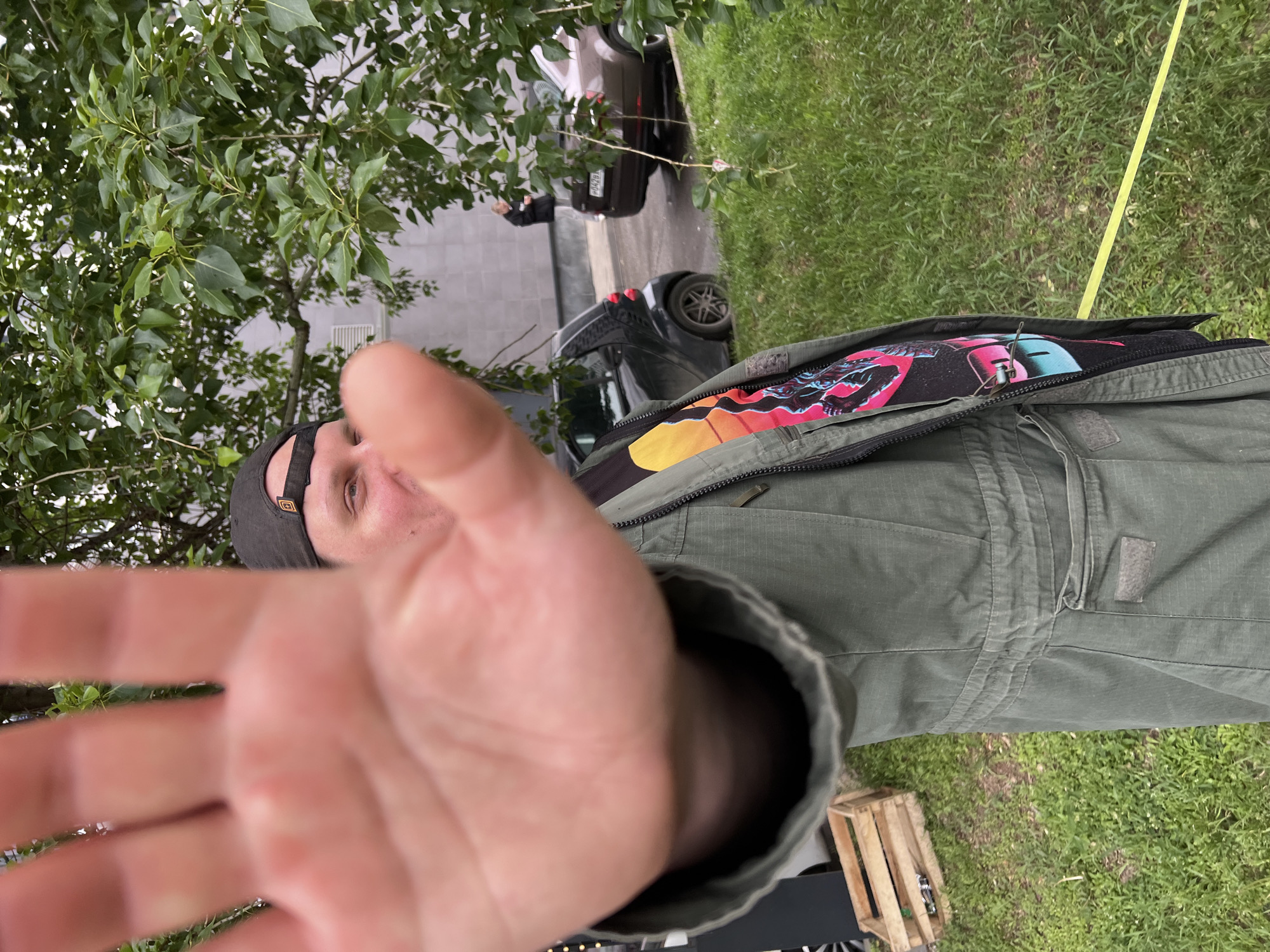 The image shows a person in an olive green jacket with a colorful t-shirt underneath, holding their hand up close to the camera, blurring the view. The person is wearing a dark baseball cap. Behind them, there is green grass and a tree. In the background, there's a street with parked cars and a building. A person in a black jacket is standing near the cars. The image appears to be taken outdoors in an urban setting. Based on the visual cues, it's not possible to confirm the location as Mytishchi, Russia.