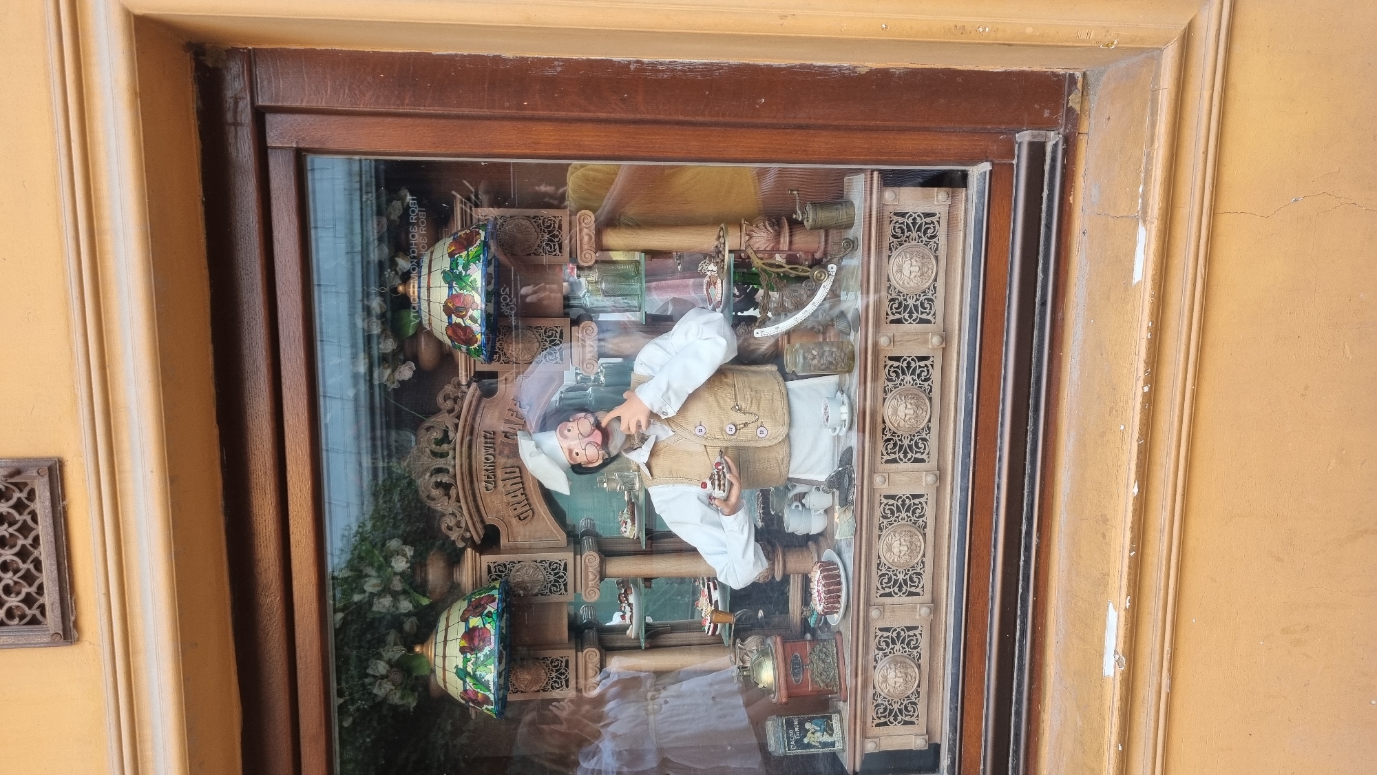 This image shows a detailed shop window display for "Czernowitz Grand Cafe" in Chernivtsi, Ukraine. The display features a mannequin dressed as a baker, wearing a chef's hat, vest, and apron. The baker is holding a small cake with red berries and appears to be contemplating it with a finger to his chin. The display includes various pastries and cakes on tiered shelves, decorative lamps with stained-glass shades, vintage coffee grinders, jars of coffee beans, and a scale. The backdrop of the display is ornate, with carved wooden elements. Reflections in the glass suggest an outdoor setting, possibly a street view, with a hint of greenery and a blurry figure in the background. The exterior of the building has a yellow facade with brown wooden window frames.