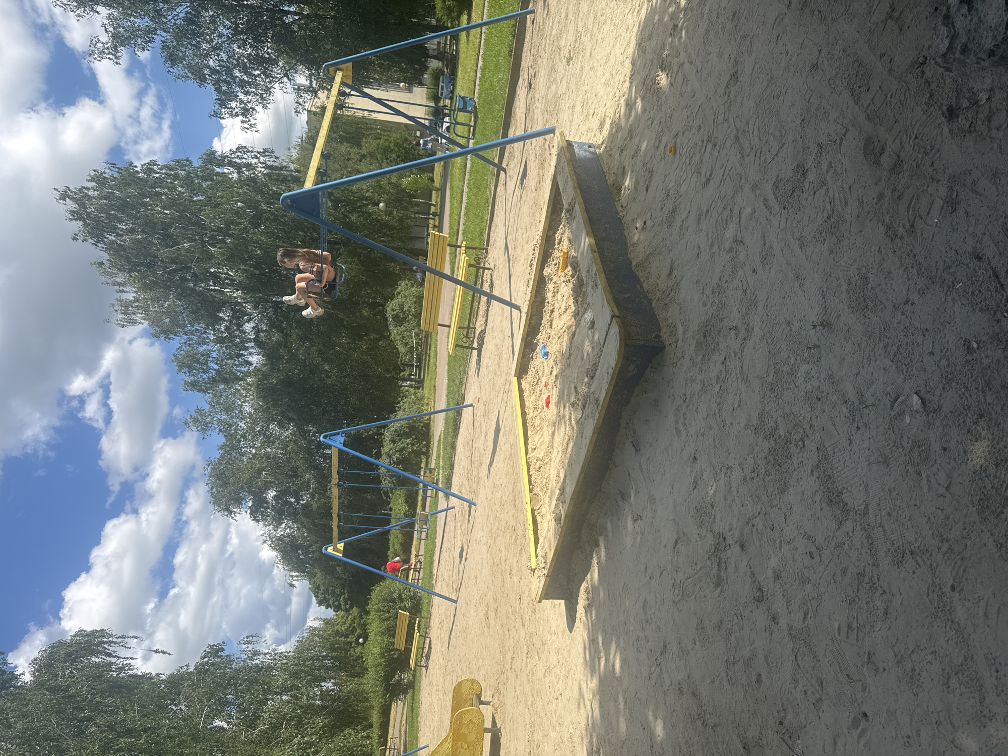 The image depicts an outdoor playground scene on a sunny day, with a blue swing set with yellow accents, a sandpit, and a yellow slide. A young girl is swinging on one of the swings, her legs extended. In the background, there are trees, benches, and buildings. The ground is covered in sand, with visible footprints. The sky is blue with scattered white clouds. The location is not specified in the image or provided in the prompt.