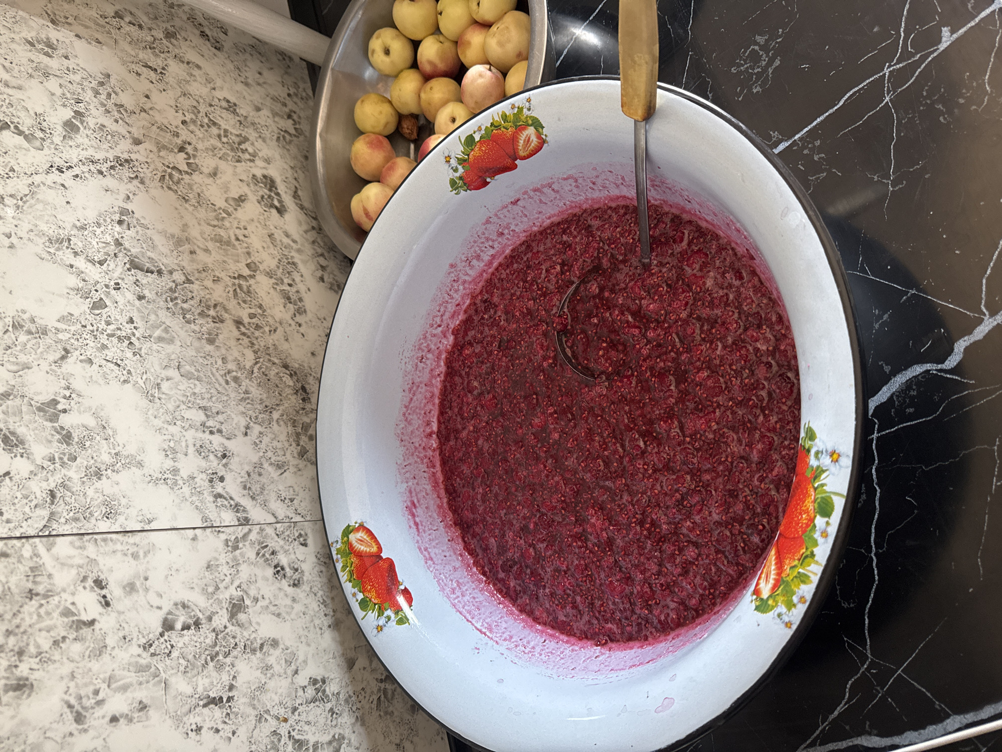 In Şardara, Kazakhstan, a large white enamel bowl filled with a bright red, mashed fruit mixture, likely raspberries, sits on a black marble countertop. A metal ladle with a wooden handle rests inside the bowl, partially submerged in the mixture. The bowl's rim is decorated with painted strawberries and small white flowers. To the right of the large bowl, a silver metal basin contains a pile of small, round, light-colored fruits, possibly apples or small plums. Behind the bowls, a grey and white marbled backsplash covers the wall. The scene appears to be a kitchen or preparation area for making jam or preserves.