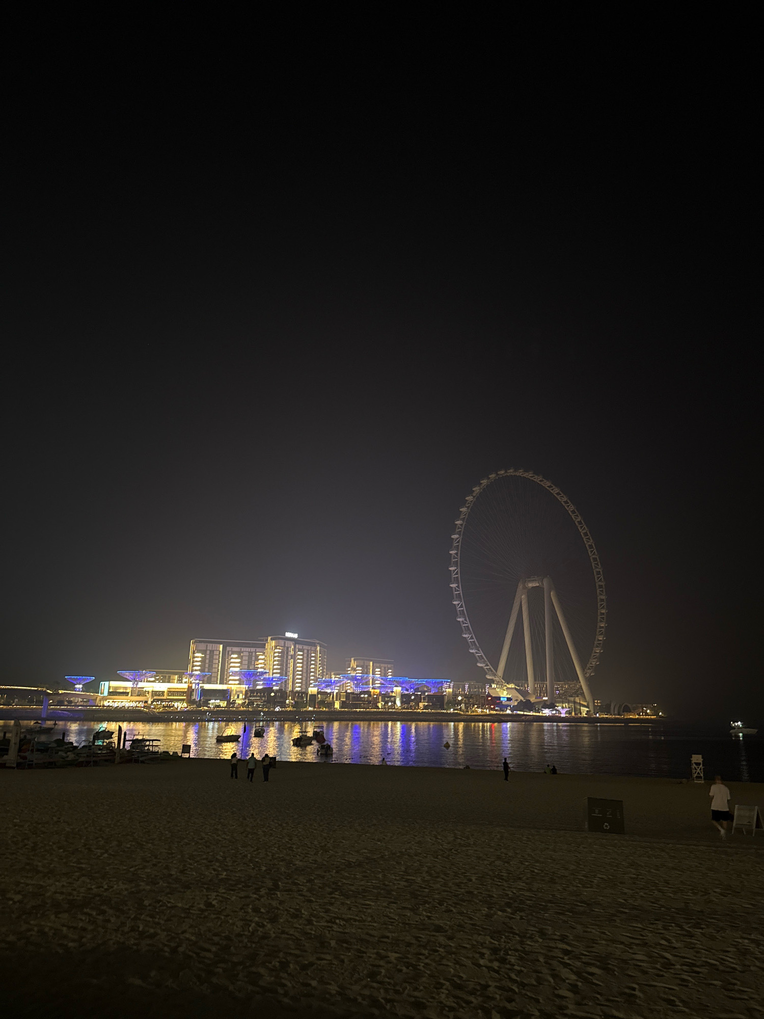 This nighttime vertical-format photograph captures a view of the Jumeirah Beach Residence (JBR) area in Dubai, United Arab Emirates, featuring the Ain Dubai observation wheel. The foreground consists of a sandy beach with subtle indentations from footprints. Three individuals, appearing as silhouettes against the illuminated background, are standing together on the sand in the mid-ground. To the left, several small boats are moored near the shore, their reflections shimmering on the water.
Dominating the mid-ground to right of the image is the Ain Dubai, an imposing observation wheel with a brightly lit white structure and a circular rim. Behind the wheel and boats, a long stretch of modern buildings lights up the horizon. These buildings are adorned with numerous windows and are illuminated by a combination of white and blue lights, creating a vibrant urban glow. The water in front of the buildings reflects this artificial light. The sky above is dark and hazy, obscuring any stars. In the far right of the mid-ground, a person in a white shirt and dark shorts is walking away from the camera, and further right, a white lifeguard chair stands on the sand. A dark sign with white text is positioned on the sand near the center-right.