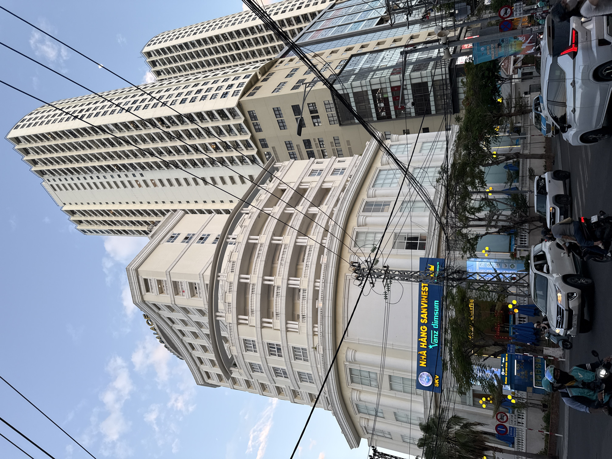 This image captures a street scene in Nha Trang, Vietnam, focusing on a curved, cream-colored building with a prominent sign reading "NHÀ HÀNG SANVINECST Vanz dimsum." Behind this building, two tall, modern skyscrapers dominate the background. The foreground shows a busy street with several vehicles, including motorcycles and cars. People are visible riding motorcycles and walking. Overhead, a tangle of power lines crisscrosses the frame, a common sight in urban Vietnamese landscapes. The sky is partly cloudy, suggesting it might be late afternoon or early evening. The architecture of the cream-colored building features a series of balconies with ornate railings and columns. The modern skyscrapers exhibit a repetitive pattern of windows. The overall impression is one of urban density and activity.
