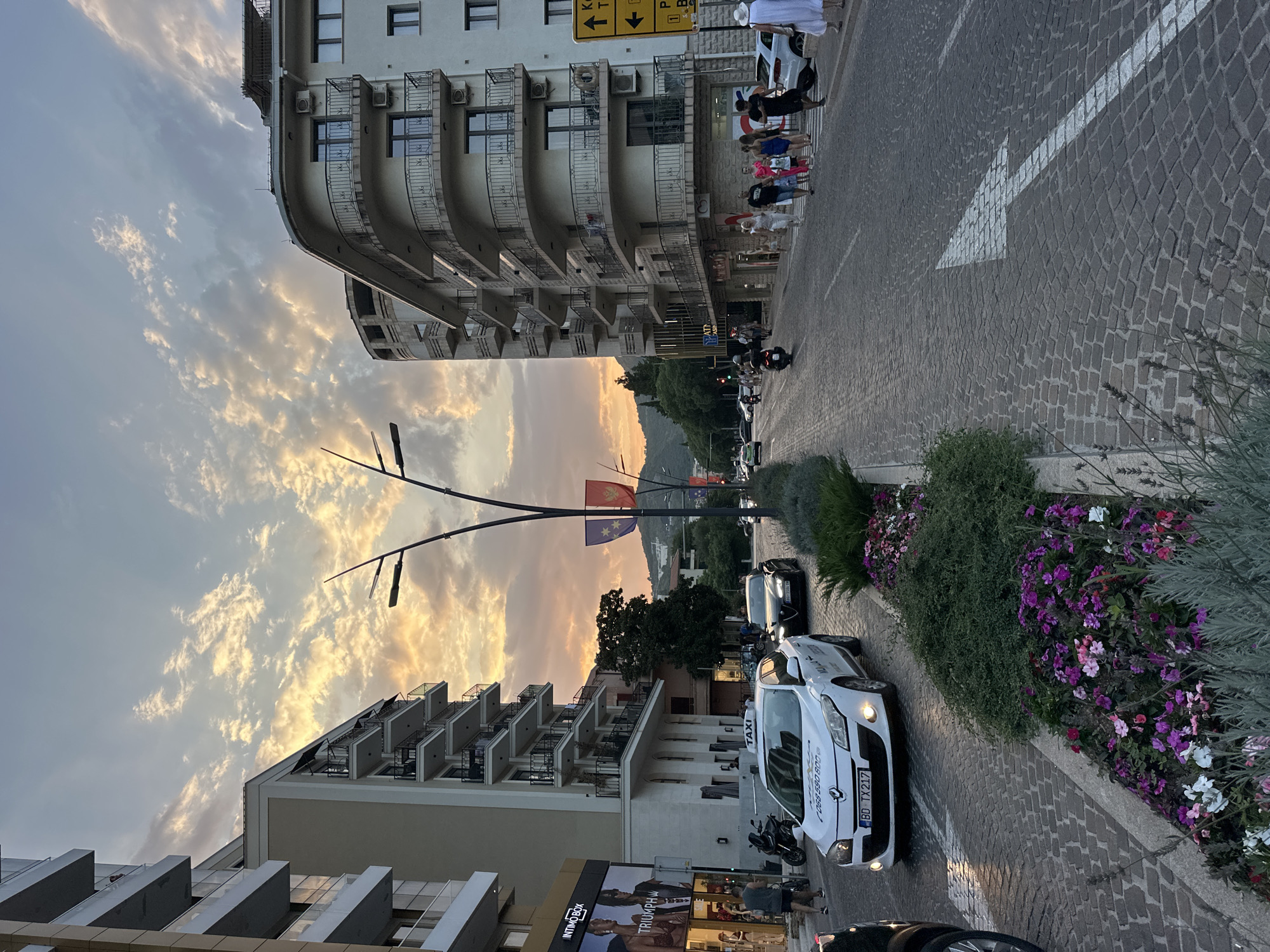 The image captures a street scene in Budva, Montenegro, at sunset. The sky is filled with dramatic clouds tinged with orange and yellow hues. Two modern, multi-story buildings with balconies line the street on either side. The street itself is paved with cobblestones and features a central median planted with various flowers and greenery.
Several vehicles are present, including a white taxi with a visible license plate "BD TX 217" and a black car in traffic. Pedestrians are walking along the sidewalk, some appearing to be tourists. Streetlights with an artistic design are mounted on poles. Flags, possibly national or regional, are displayed on poles. The overall atmosphere suggests a lively urban environment during the evening.