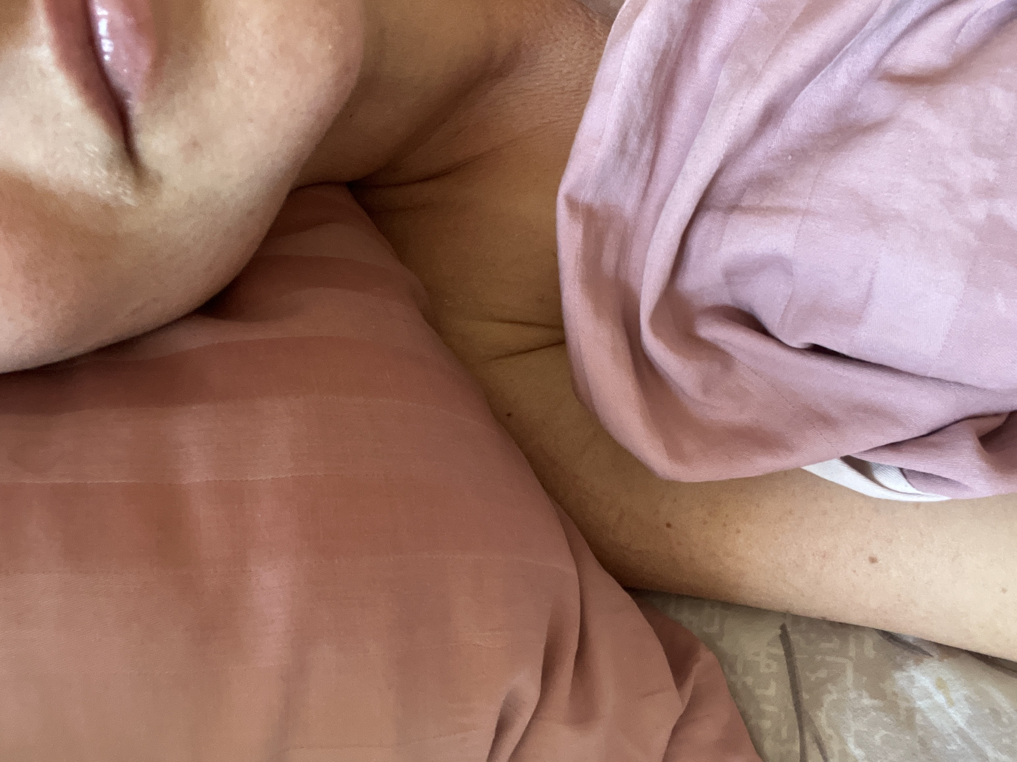 A close-up, over-the-shoulder shot shows a person's upper chest, neck, and mouth, partially covered by pink bedding. The person is lying down in bed. The scene is likely indoors, and there is no specific indication of location within Novosibirsk, Russia.