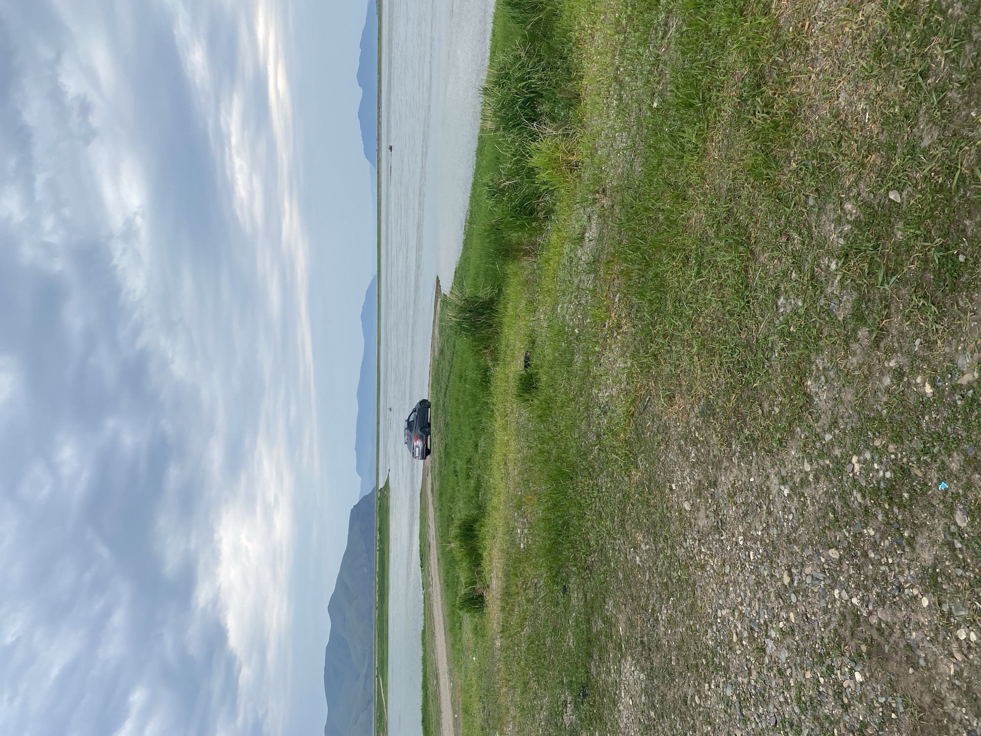 A dark grey sedan with a roof rack is parked on a dirt patch next to a wide, calm body of water. The car has a red and white sticker on its rear windshield. The foreground is dominated by a grassy embankment with a gravel path leading down towards the water. In the background, rolling, green mountains rise against a cloudy, overcast sky. The water extends to the horizon where it meets more distant, muted mountain ranges. The scene appears to be a natural, undeveloped area.