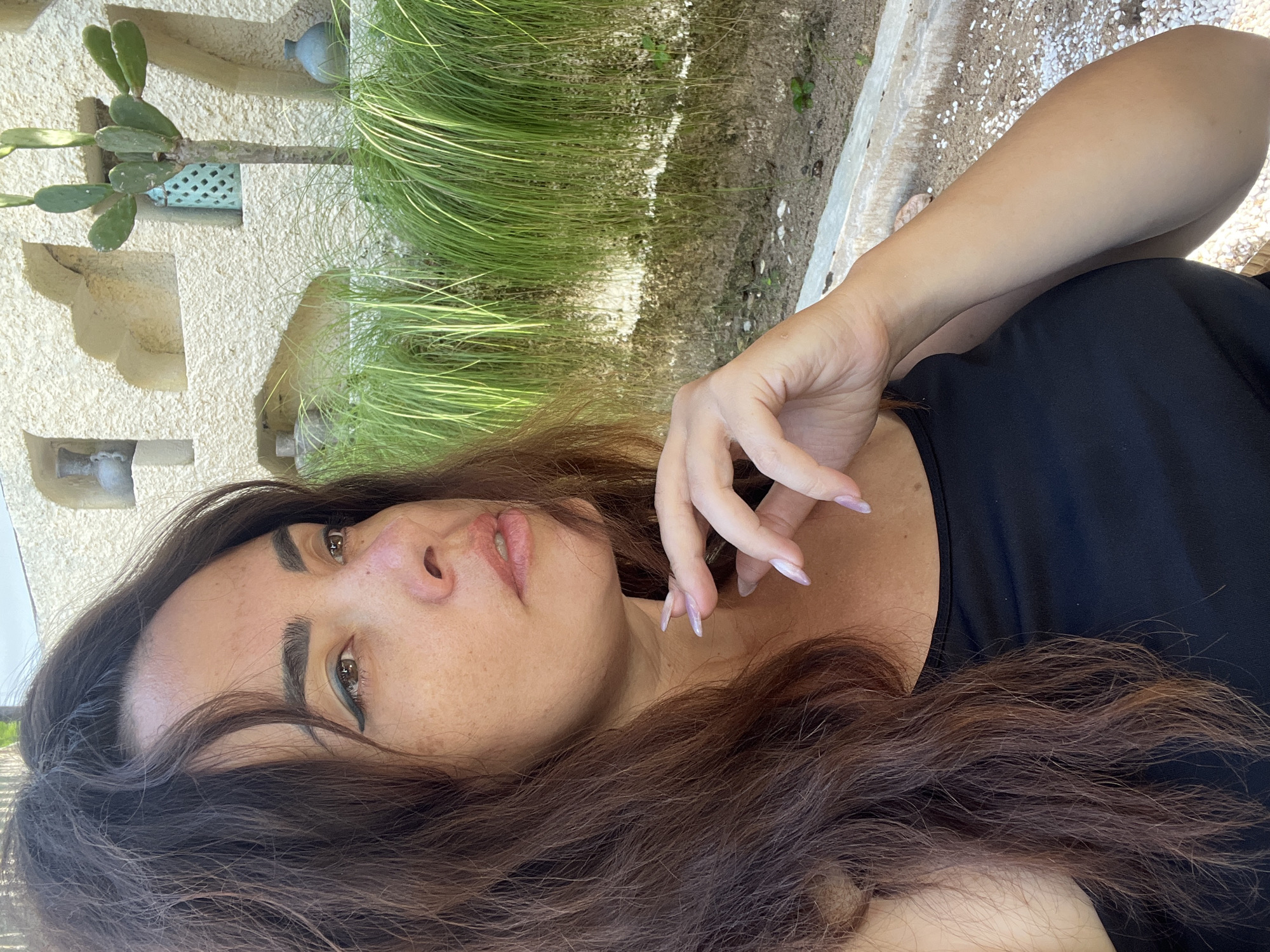 A woman with long, brown, wavy hair is shown in a close-up shot, looking to her right with a thoughtful expression. She is wearing a black tank top. Her hand, with light pink nail polish, is gently resting near her chin, with her index finger extended. The background features a textured, pale wall with decorative niches, a cactus plant, and lush green grass. The setting appears to be outdoors, possibly in Kuta Selatan, Indonesia, given the architectural style and vegetation.
