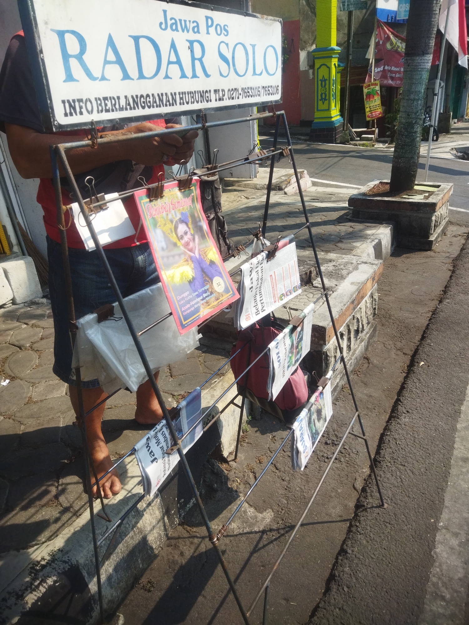 The image depicts a street scene in Giripurwo, Indonesia, focused on a man selling newspapers from a rack. The sign above him reads "Jawa Pos RADAR SOLO INFO BERLANGGANAN HUBUNGI TLP. 0271-7653105/7653106". He is wearing a red t-shirt and denim shorts, with his bare feet visible. The newspaper rack is made of metal and holds several newspapers, some of which are clipped open. In the background, there are buildings with colorful accents, a palm tree in a planter, and a street. The ground is paved with cobblestones and asphalt. The lighting suggests it is daytime, with shadows cast by the man and the rack.
