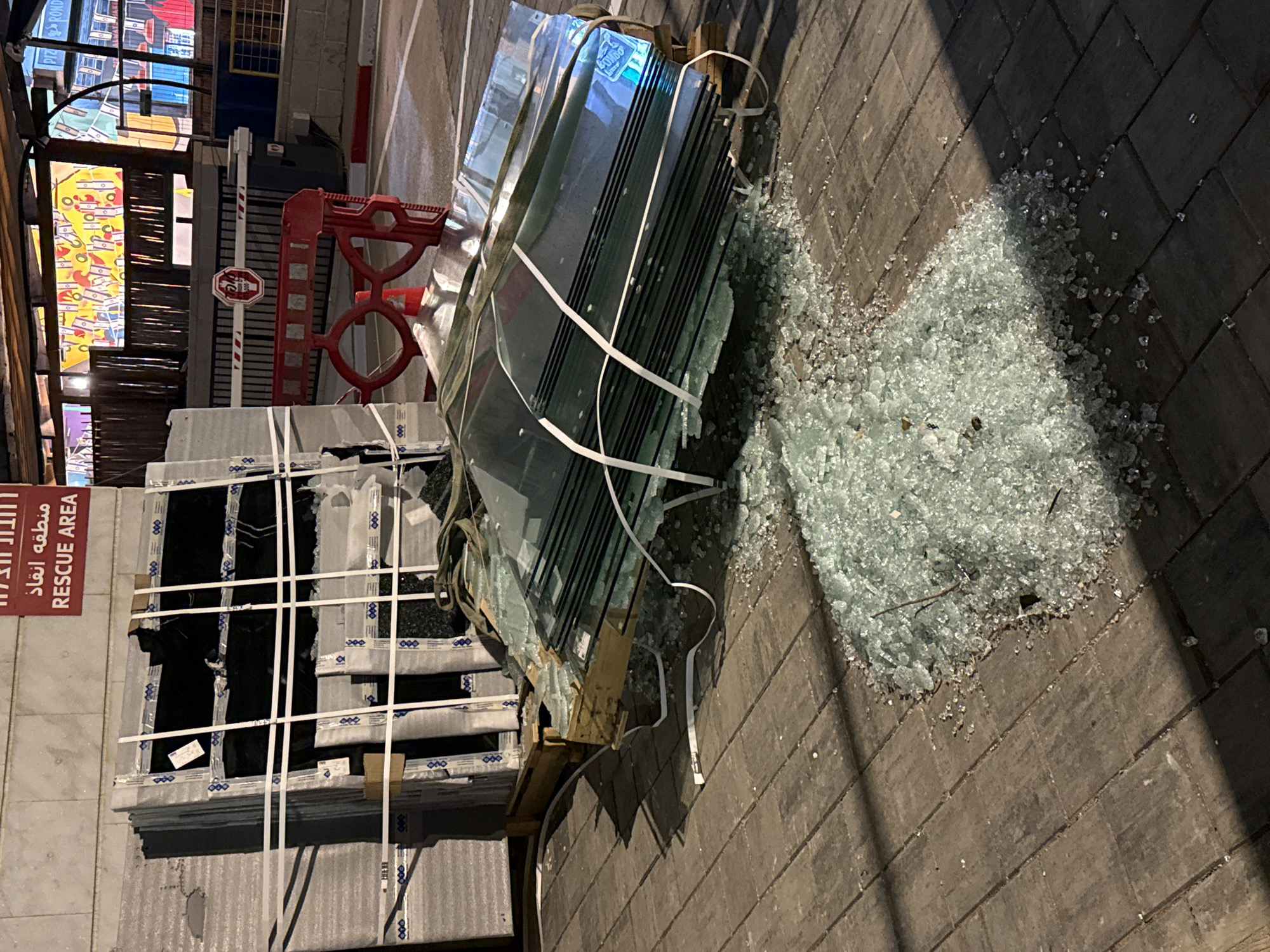 The image shows a stack of glass panes on a wooden pallet, some of which are broken, with shattered glass scattered on the brick pavement. The pallet is leaning against another stack of boxed materials, possibly windows. A sign in Hebrew and English reads "RESCUE AREA" is visible next to the boxed material. In the background, urban features include buildings, a red barrier, and lighting. The scene is likely in Petah Tikva, Israel, based on the signage.