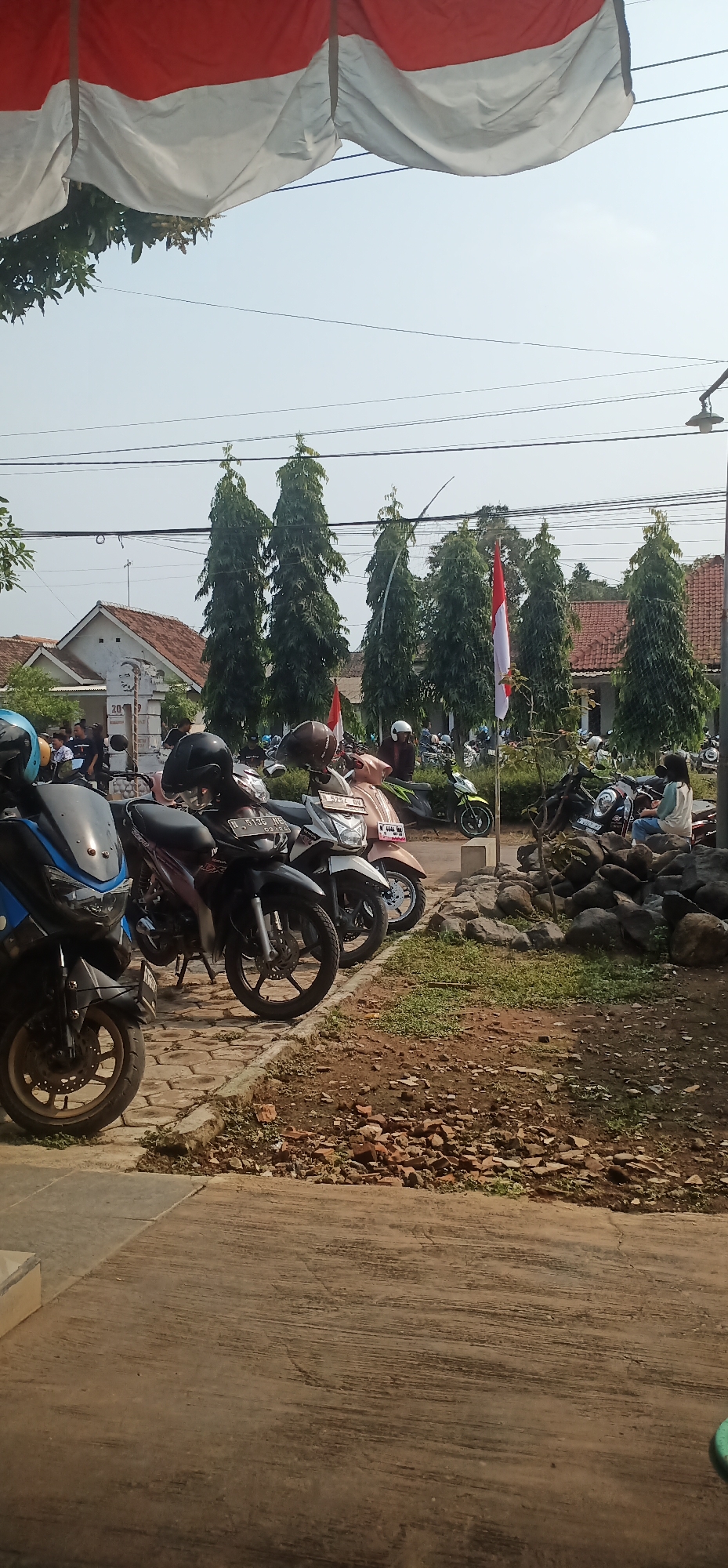 This is a vertical, eye-level, medium shot photograph of an outdoor scene in the city of Sumberkerang, Indonesia, under a clear sky. In the foreground, a tiled walkway leads to a paved area where several parked motorcycles are visible. To the left, a blue and black motorcycle is partially visible. Adjacent to it, a black motorcycle with a maroon seat and a red license plate (N 5136 HE) is parked. Another white scooter with a pink seat and a black license plate (N 4757 BV) is positioned behind it, followed by a lime green scooter. Further back, more motorcycles are parked, some with riders.
The middle ground features a line of tall, slender trees. Behind the trees, a building with a red tiled roof and white walls is visible. In front of the building, there appears to be a monument or statue. Two Indonesian flags, red and white, are planted in the ground among the trees.
In the background, a few more buildings with red roofs are scattered. Electrical wires run across the top of the frame. A large red and white banner, possibly a flag or decoration, is draped across the top of the image. The overall scene suggests a public gathering or event in a residential or community area. The ground is covered in dirt, scattered rocks, and some dry leaves. The lighting indicates it is daytime.