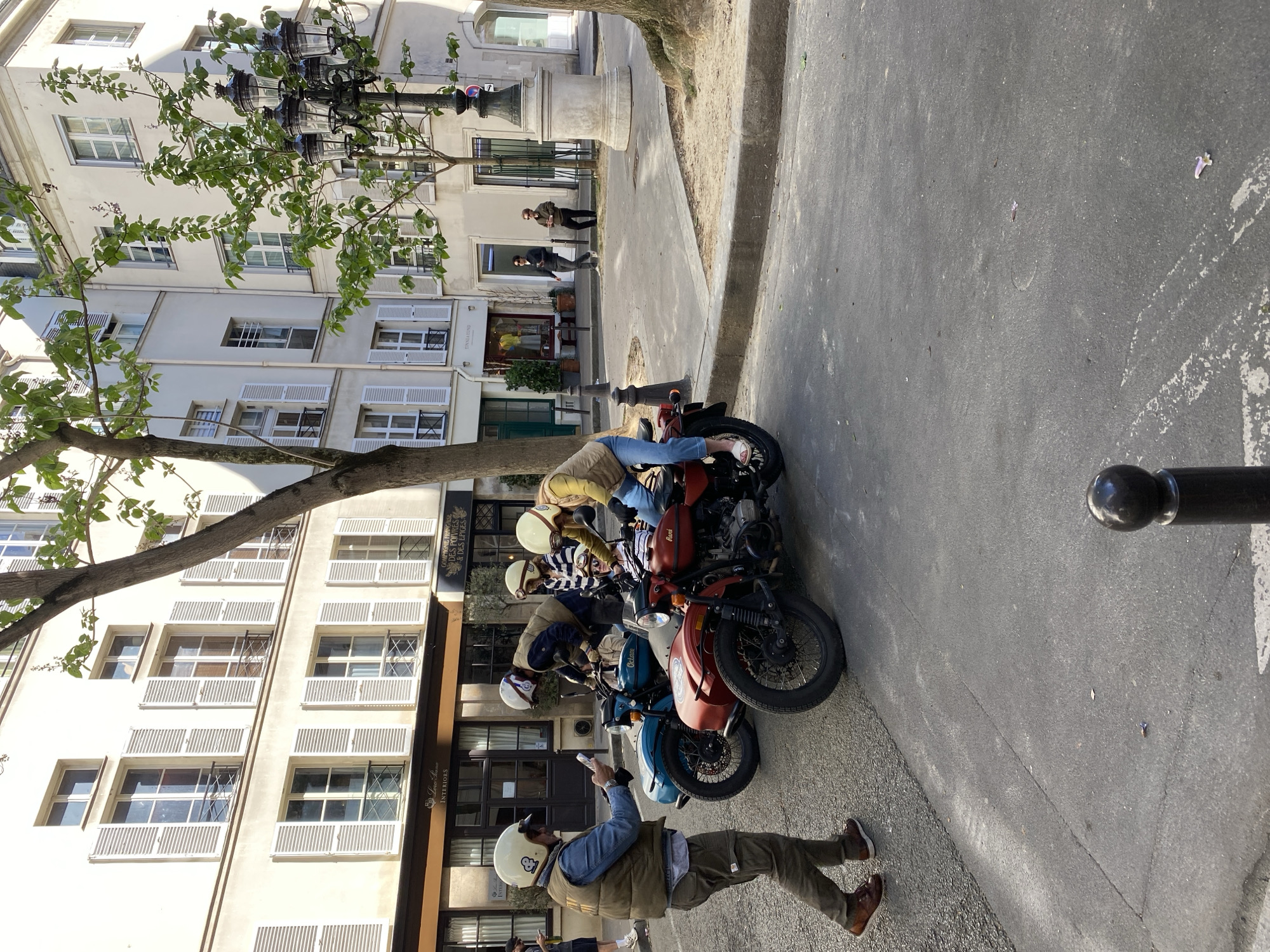A bright, sunny street scene in Paris, France, captures a casual gathering of motorcyclists and their classic-style bikes. The image is taken on a clear day, evidenced by strong shadows and vibrant lighting on the pavement and buildings.
In the foreground, a person wearing a white helmet, a brown vest over a blue denim jacket, olive green pants, and brown shoes stands on the pavement. They are holding a phone up, appearing to capture a photo or video of the group or the surroundings. To their left, a line of retro-style motorcycles is parked, including prominent red and blue models, with other bikes partially visible. Several other individuals, also sporting helmets and casual jackets or vests, are either seated on or standing near these motorcycles, suggesting a group outing or a stop during a ride.
The setting is characterized by traditional Parisian architecture, featuring multi-story, light-colored buildings with numerous windows adorned with white louvered shutters, some open and some closed. A large tree with verdant leaves partially obscures the buildings on the left side of the frame, and a prominent street lamp with multiple dark fixtures is visible among its branches. Further down the street, near a stone pillar or fountain structure, two distant figures can be seen.
Visible text on a building facade reads "DÉCORS DES POUTRES & DES ÉPICES" and below it, "Luxe du homme INTERIORS," indicating commercial establishments. The overall impression is one of relaxed camaraderie and a shared enjoyment of the urban environment.