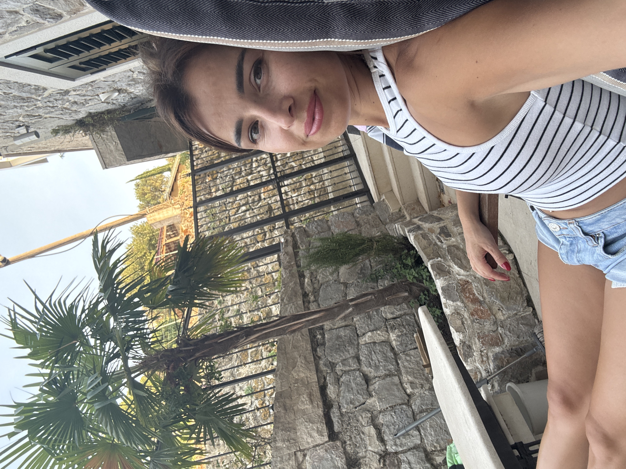 A woman with brown hair and fair skin is sitting in an outdoor chair, looking at the camera with a slight smile. She is wearing a white tank top with black stripes and blue denim shorts. Her legs are crossed and extended. Behind her, a stone wall, a palm tree, and a staircase are visible. The setting appears to be a courtyard or patio, possibly in a Mediterranean climate.