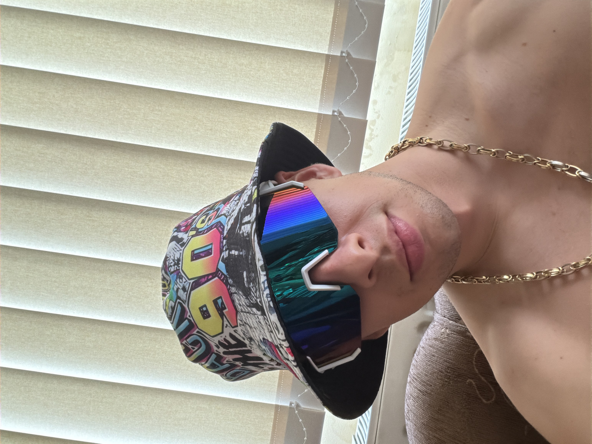 A person wearing a bucket hat, sunglasses, and a gold chain is posing in front of a window covered with blinds. The individual is facing the camera with their head tilted to the left. The bucket hat has a black brim and a multicolored pattern on the crown. The sunglasses have iridescent lenses and white frames. The gold chain is visible around the person's neck. The environment appears to be indoors, with blinds covering the window. The location is Unknown, Unknown.