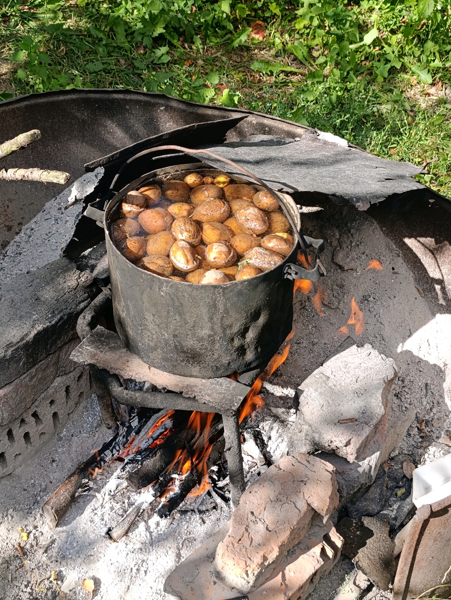 In Chereshneve, Ukraine, a large metal pot filled with potatoes and water is being heated over an open fire. The pot rests on a makeshift grate fashioned from metal bars, which is supported by a ring of bricks or stones. Embers and flames are visible beneath the pot, indicating the fire is active. The surrounding area appears to be outdoors, with dry grass and vegetation visible in the background. Scattered stones and ash suggest a rustic, possibly temporary, cooking setup. The scene is captured from a high angle, looking down into the pot and the fire.