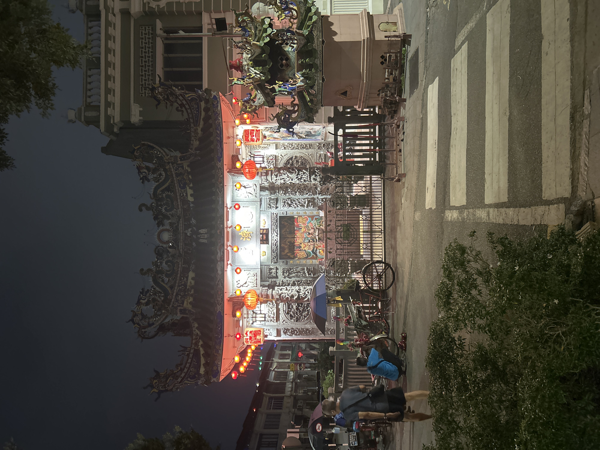 The image depicts a scene at night in George Town, Malaysia, in front of a temple. The temple features ornate architecture with intricate dragon carvings on its roof and walls, illuminated by numerous red lanterns. In the foreground, a pedestrian crossing is visible on the asphalt road. To the left, two individuals are seen. One person, wearing a dark t-shirt and shorts, is walking away from the camera. Another person, in a blue shirt, is crouched down near a bicycle. Several bicycles are parked nearby, along with two open umbrellas, one blue and one black. A decorative structure, resembling a miniature temple, stands to the right of the main temple entrance. The overall environment suggests a street scene in a historic urban area.