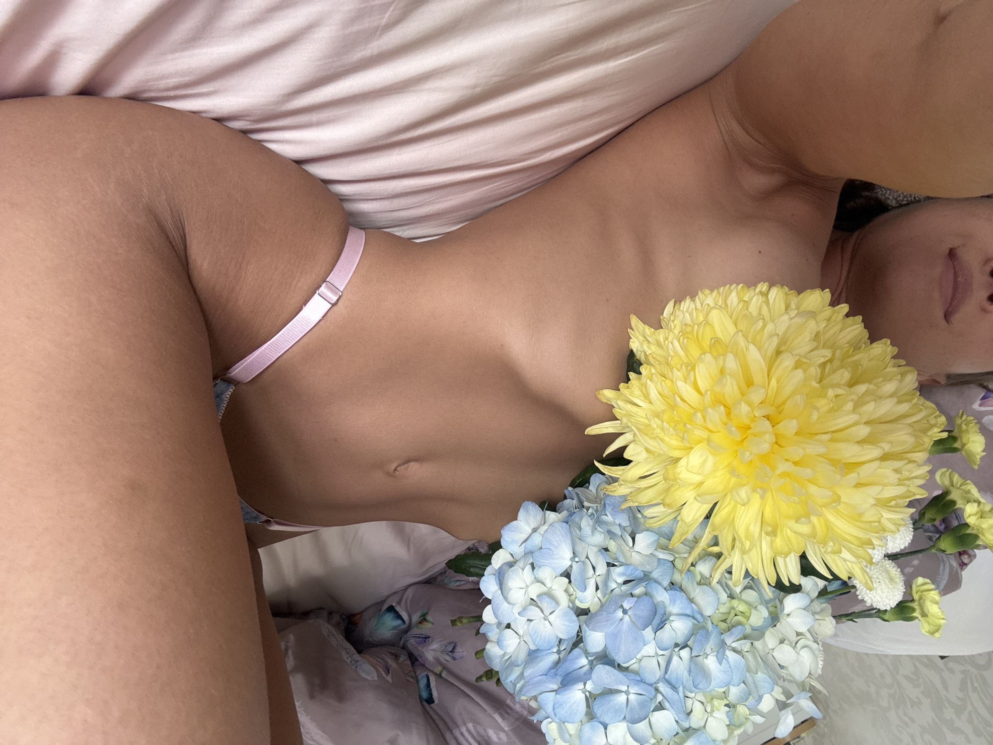 A woman is lying on a bed covered with a pink satin sheet. She is partially nude, wearing a pink bikini bottom. A bouquet of flowers, including a large yellow chrysanthemum and blue hydrangeas, is placed near her chest. The background consists of bedding with a floral pattern. No specific location in Saint Petersburg, Russia, can be identified from this image.
