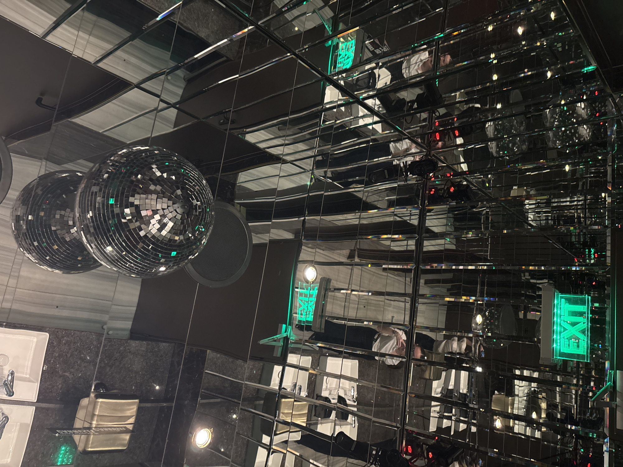The image depicts the interior of a bathroom, likely a public restroom, possibly located in Sofia, Bulgaria. The ceiling and walls are covered in square mirrors, creating an infinite reflection effect. Two disco balls hang from the ceiling. There are sinks, a soap dispenser, and an EXIT sign. Reflections of people can be seen in the mirrors.