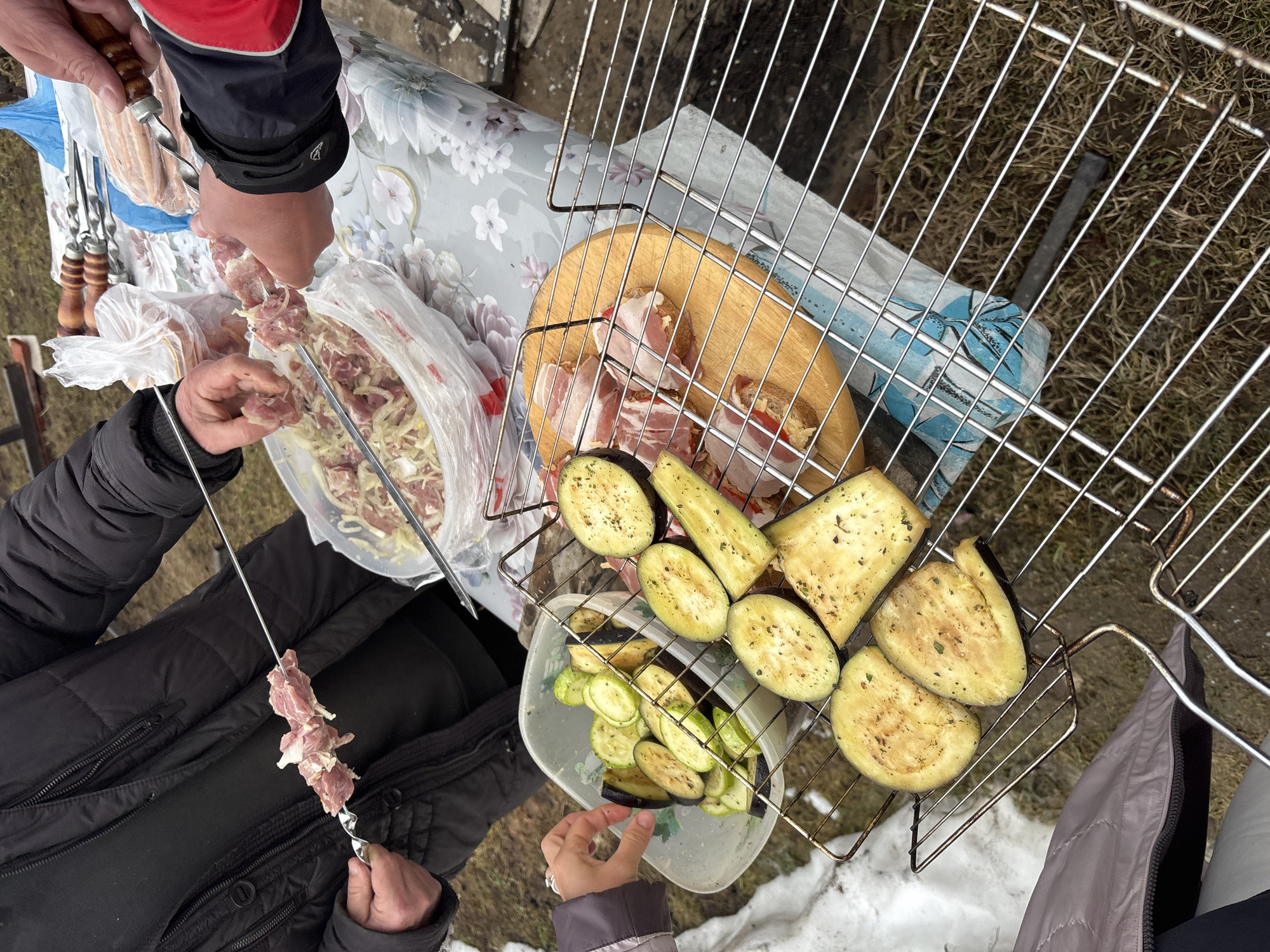 The image depicts a scene outdoors, likely in a backyard or picnic area based on the presence of snow on the ground. Two people are preparing food for a barbecue. One person, wearing a jacket with red and black sleeves, is skewering meat from a plastic bag containing meat mixed with onions. Another person, in a dark puffy jacket, also holds a skewer with meat.
A metal grill sits on top of a table. On the grill, there are several items being prepared: grilled eggplant slices, grilled zucchini slices in a plastic container, and bread slices wrapped in bacon on a wooden cutting board.
The environment appears to be a residential area, with a glimpse of a building in the background. The image suggests a communal meal preparation taking place in a casual, outdoor setting. The location is in the city of Chaikovsky, Russia.