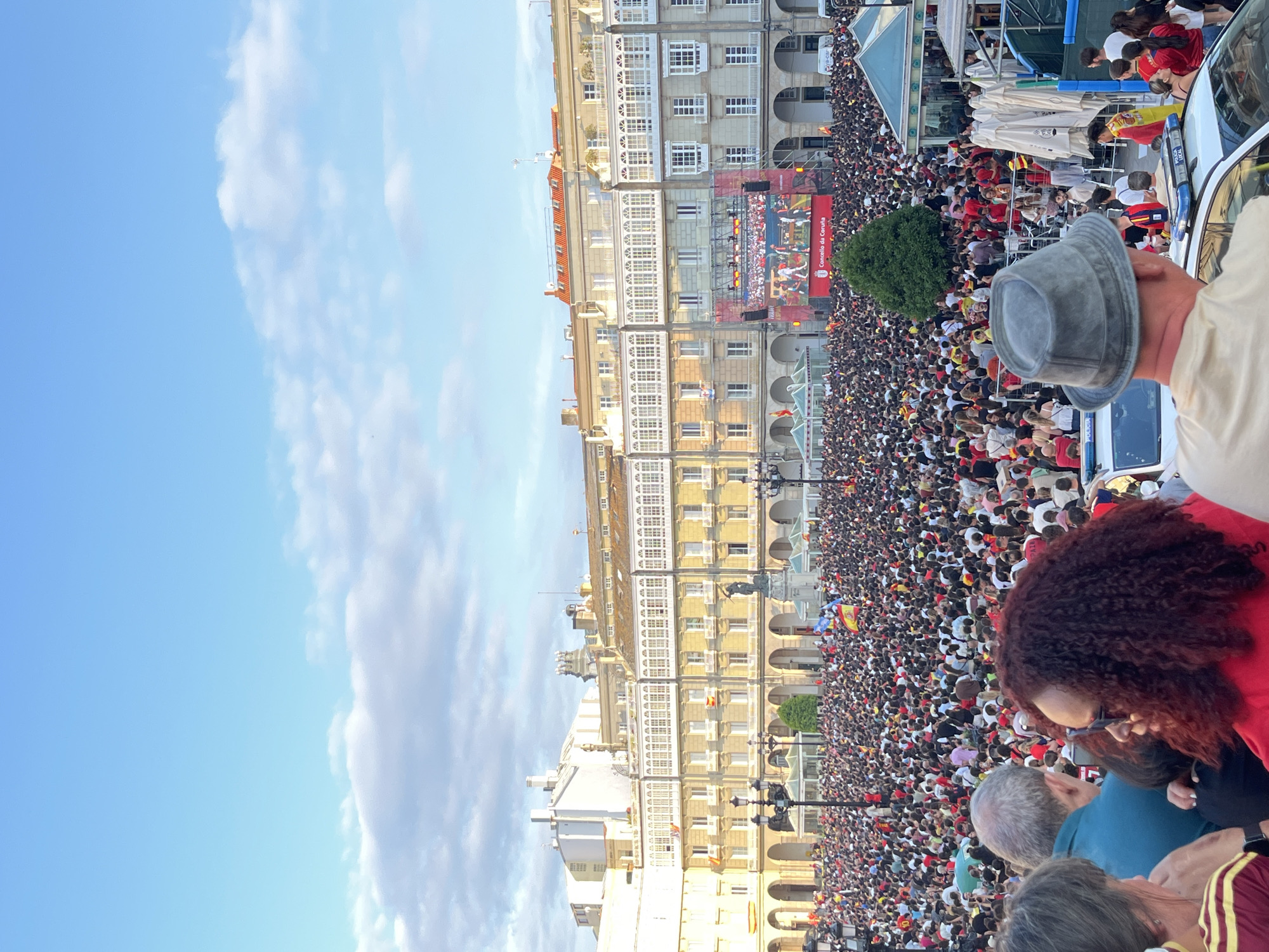 The image shows a large outdoor gathering of people in a city square, likely for a public viewing of an event, given the large screen displaying images of what appears to be a sporting event. The screen is set up in front of a row of multi-story buildings with distinctive white balconies. A significant crowd is packed into the square, with many individuals wearing red and yellow, colors associated with Spain. Some people are waving Spanish flags. The architecture suggests a European city, and the presence of "Concello da Coruña" on the screen confirms the location as A Coruña, Spain. The sky above is blue with scattered white clouds, and the lighting suggests it is late afternoon or early evening. In the foreground, there are police vehicles with flashing lights, indicating a managed public event. A few individuals are more clearly visible in the foreground, some looking towards the screen, others interacting amongst themselves. The overall atmosphere appears to be one of a communal celebration or viewing event.